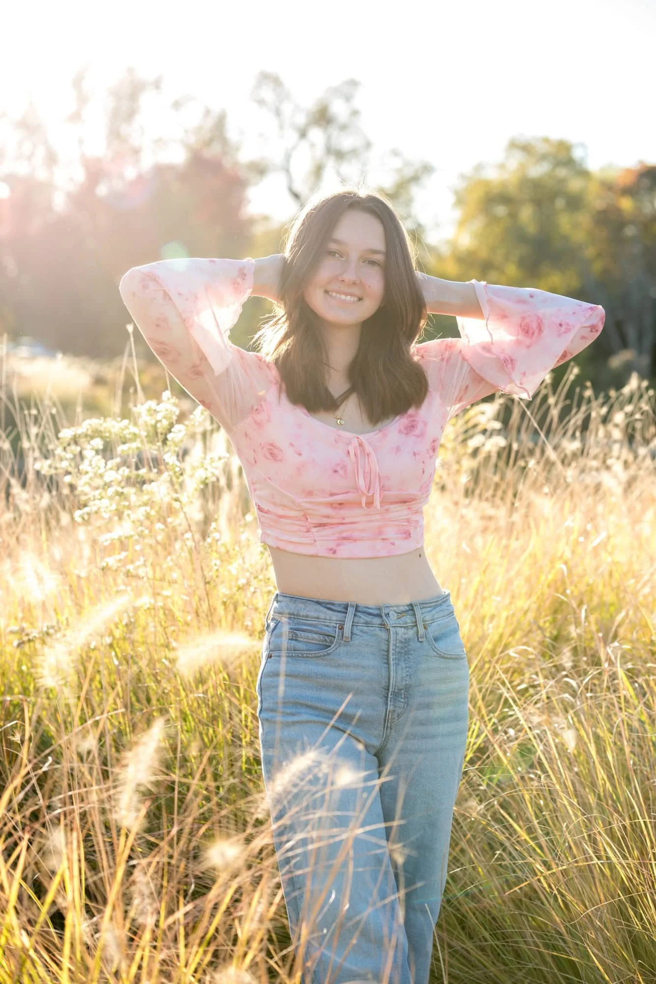 In a field of tall ornamental grasses by the Nathan Frank Pagoda, a senior girl stands in a pink floral crop top and blue jeans with her hands behind her head during a whimsical style photo shoot.