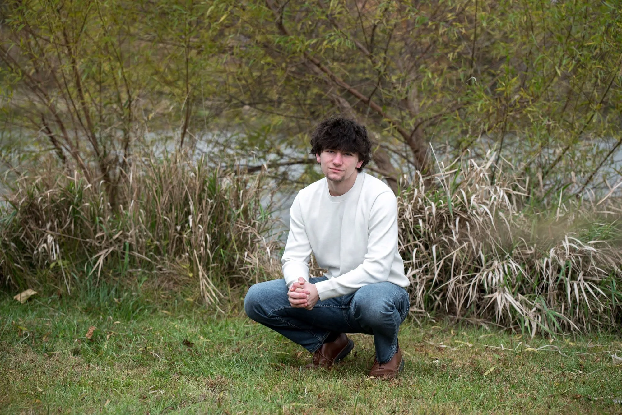 Squatting in the grass near a brushy shoreline, a senior guy smiles during a fall session with a Defiance photographer.
