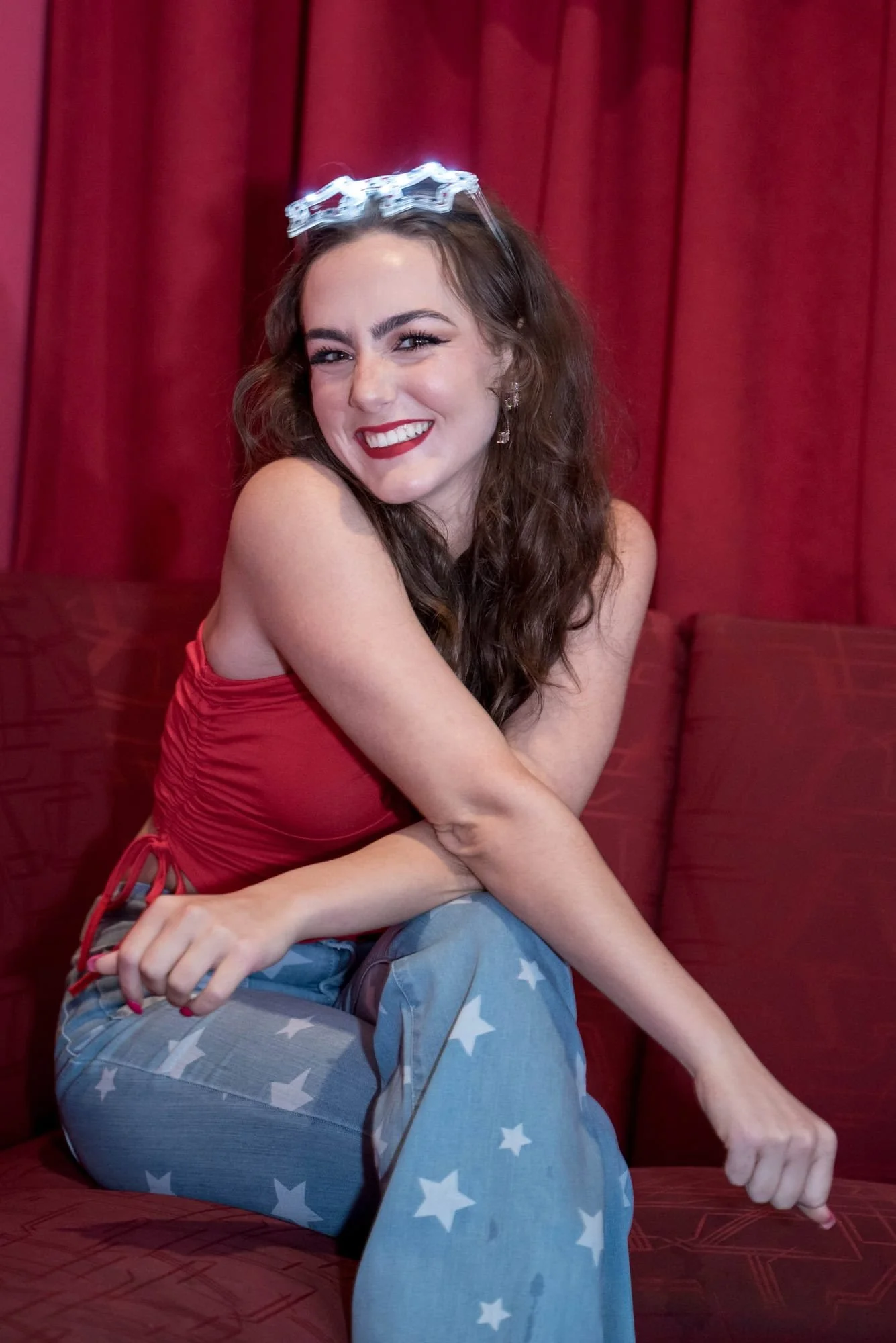 Sitting on a couch inside the Pin-Up Bowl venue, a senior girl in a red tank top and star-patterned blue jeans with star sunglasses poses for a picture.