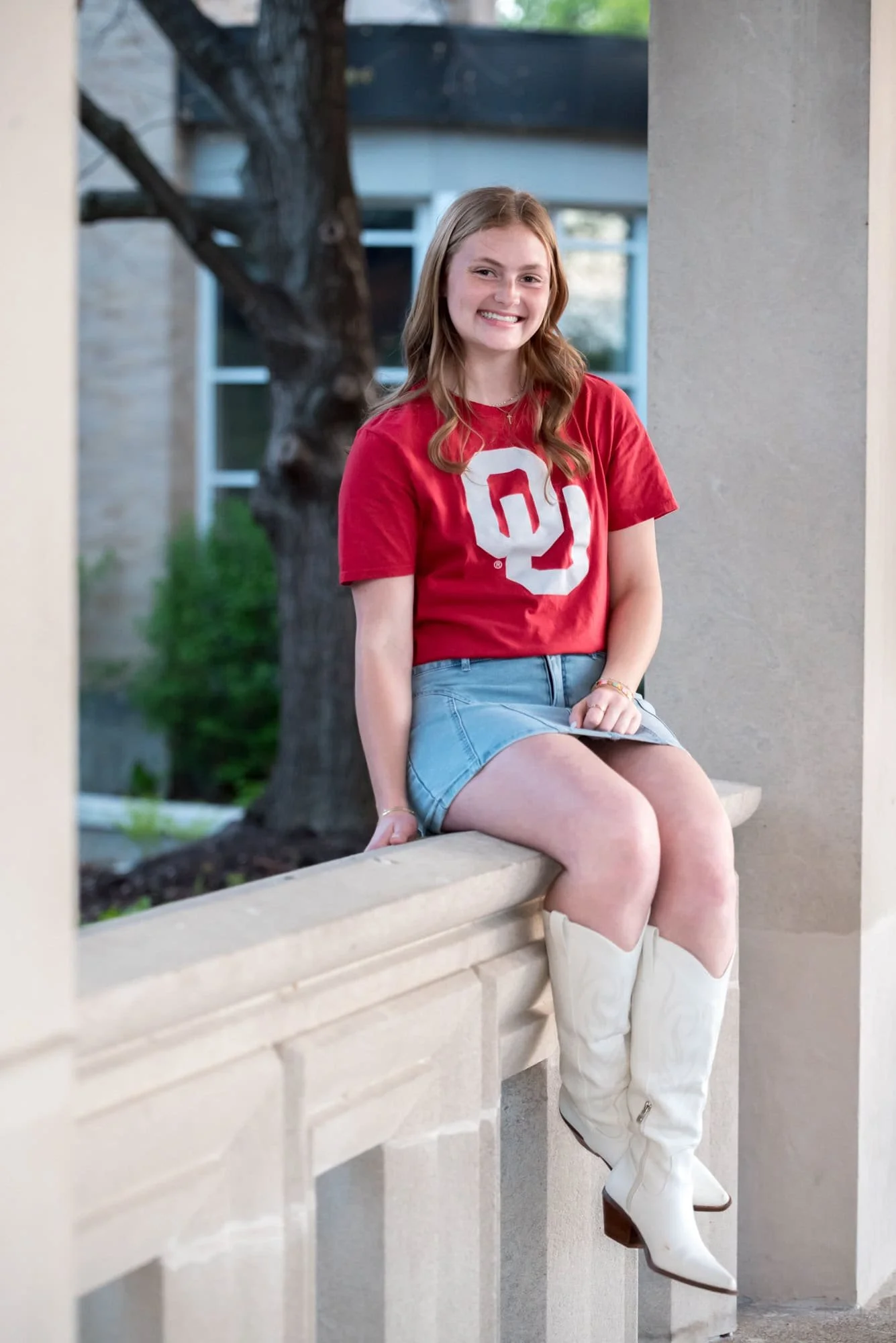 A photo shows a high school senior girl wearing a red Oklahoma University shirt, a jean skirt, and white boots at The Muny while sitting on a ledge.