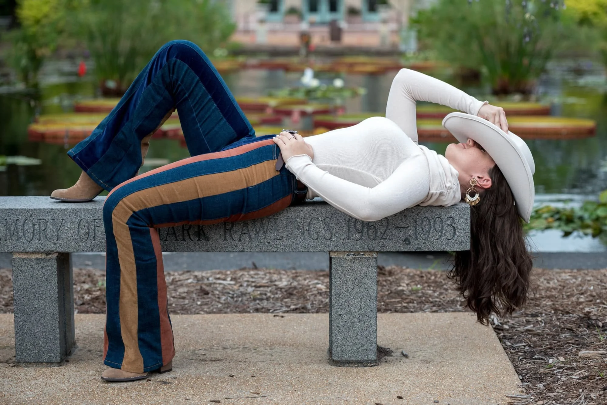 A photo shows a senior girl wearing a long-sleeve white top and striped blue jeans lying down on a bench in St. Louis County with her white cowboy hat resting over her face.