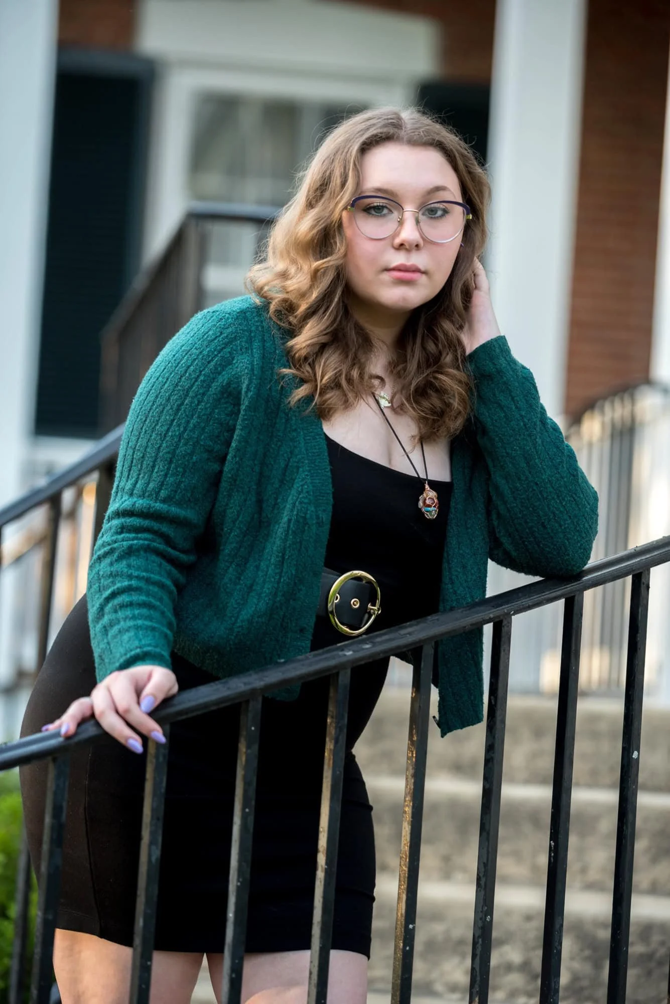 Standing on the steps of the Victorian home at Queeny Park, a senior girl in a black dress and green cardigan poses for a photo.