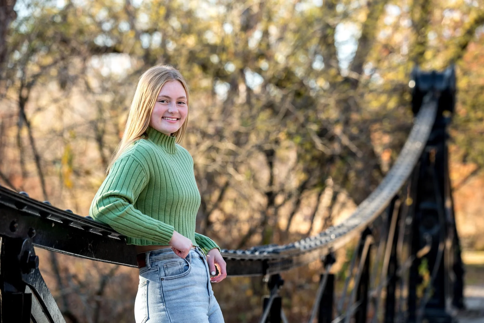 Senior girl in a green sweatshirt and jeans leaning on a black Victorian bridge during the fall at Forest Park, taken by a St. Louis senior photographer.