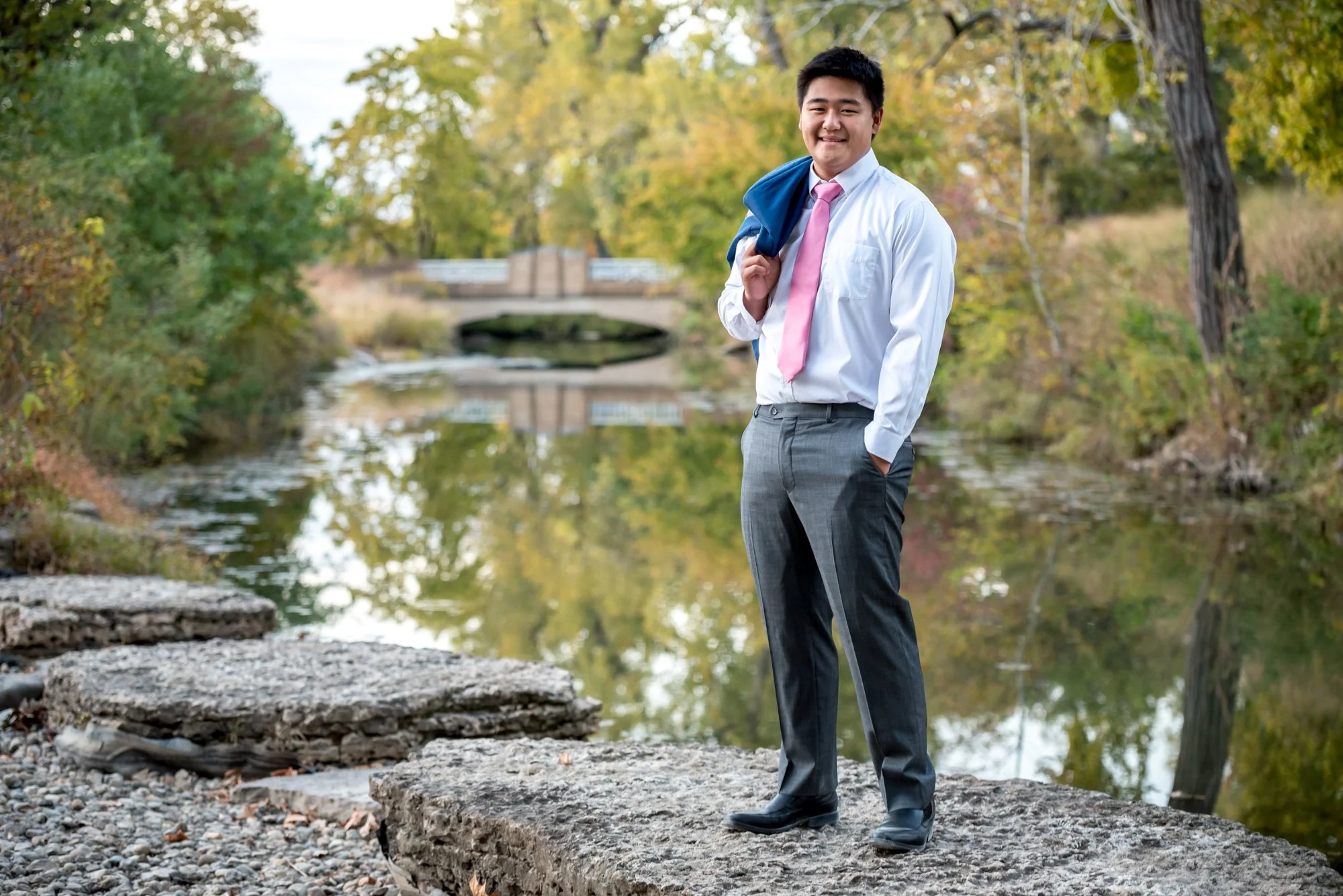 Asian senior guy wearing a white button-up shirt, pink tie, and grey suit pants, standing on a rock in front of a creek during the fall down by the Art Hill lake basin at Forest Park for his senior photo.