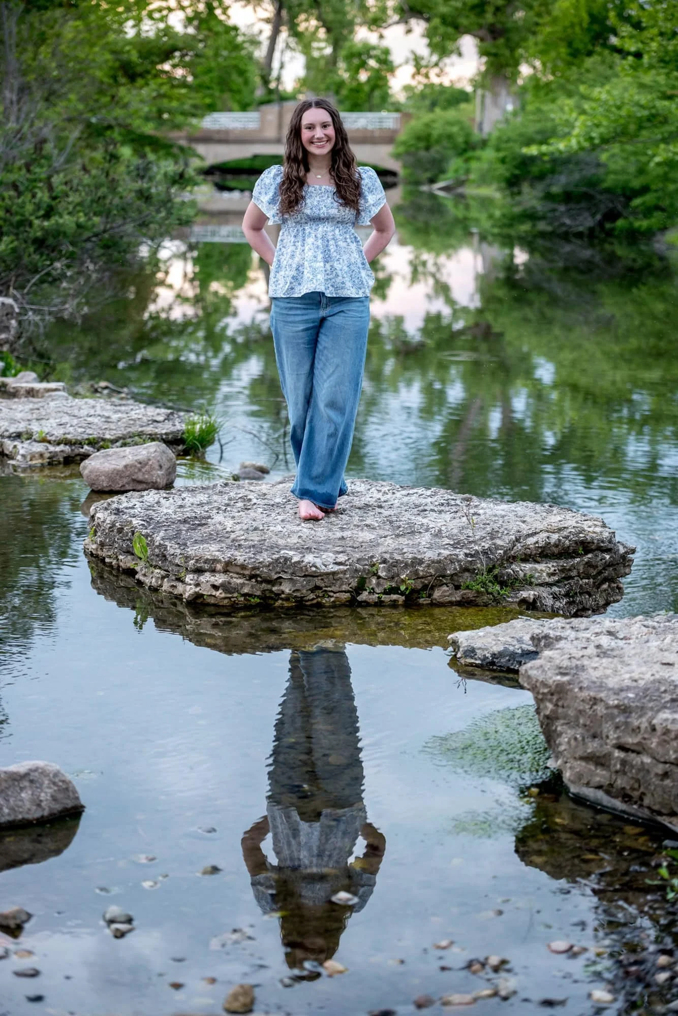 Standing on a flat stone in the water, a smiling senior girl poses as her reflection mirrors perfectly in the calm surface of a creek near Art Hill in Forest Park.