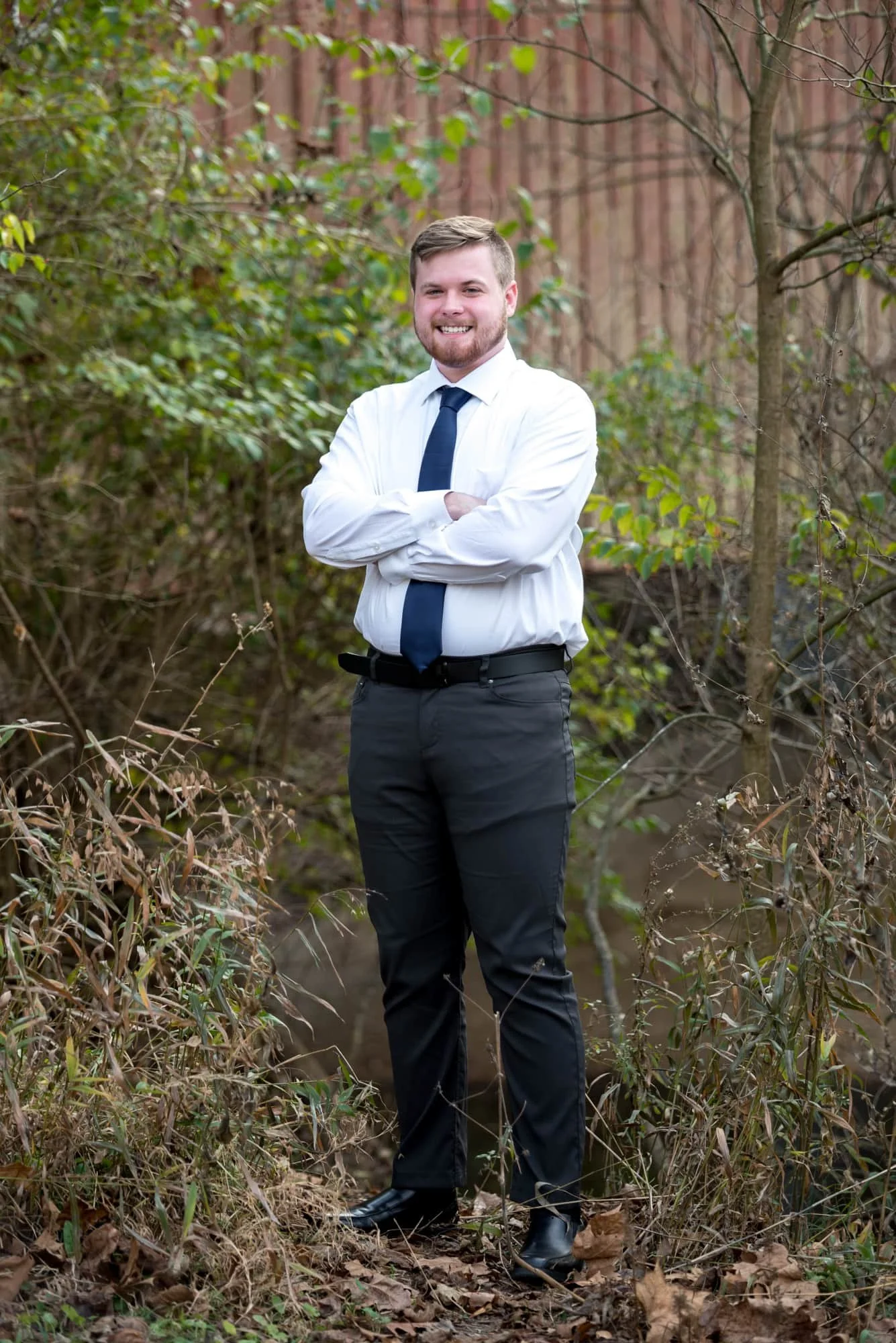 Dressed in a white button-up shirt, navy blue tie, and dress pants, a high school senior guy stands outside the red Sandy Creek Covered Bridge for a photograph.