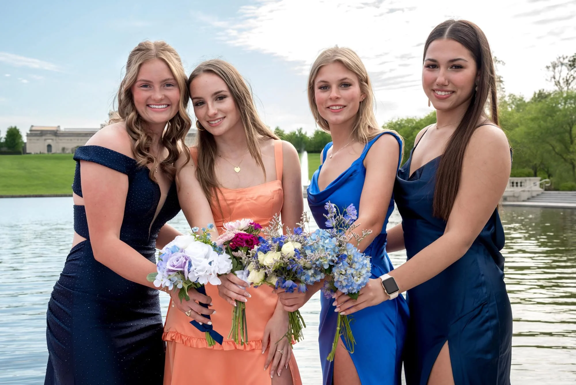 Wearing prom attire, a high school senior girl in a navy blue dress poses with three friends along the Art Hill lakeside for a photo.