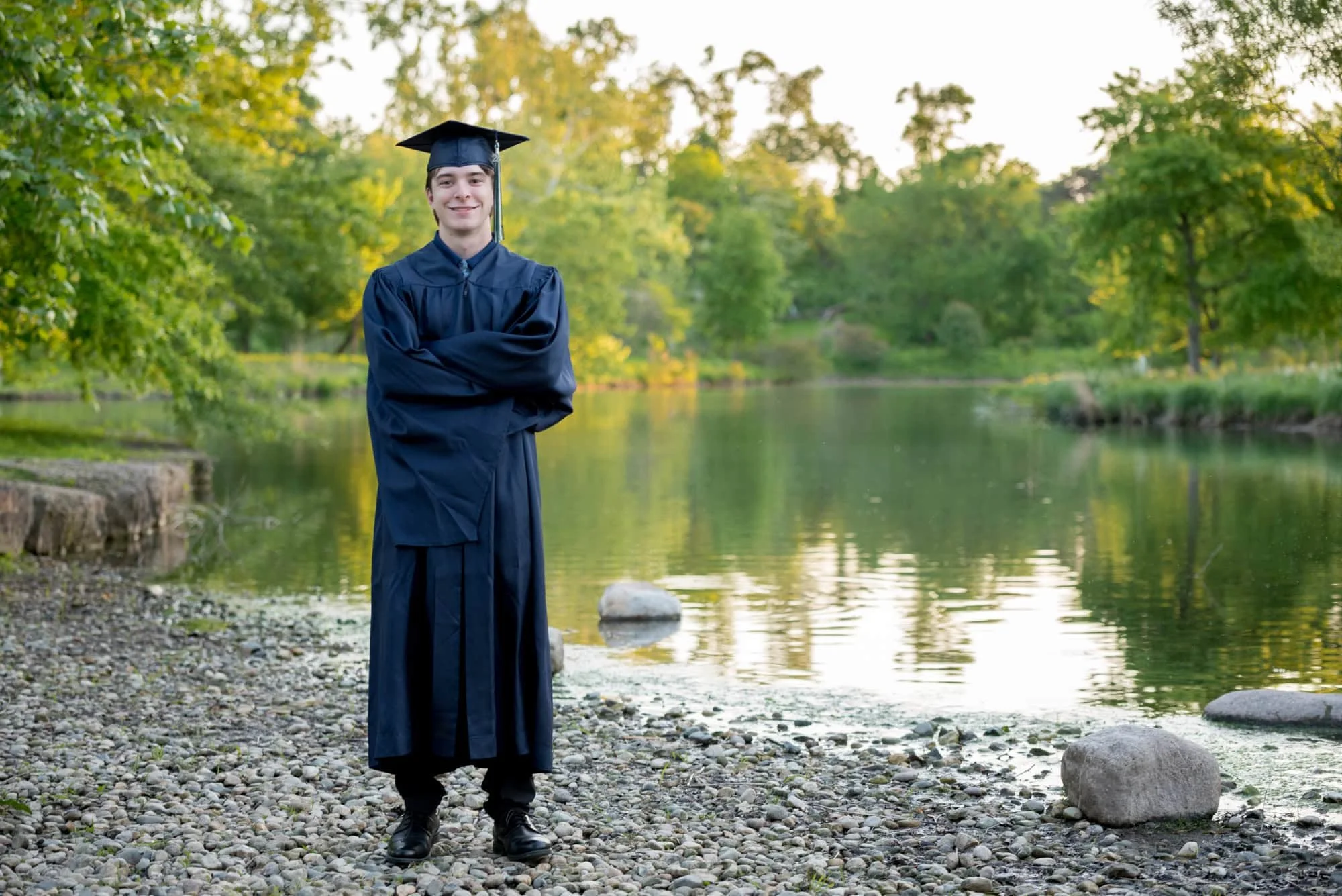 High school senior boy standing with arms crossed in his graduation cap and gown on a rocky creek bank at Forest Park.