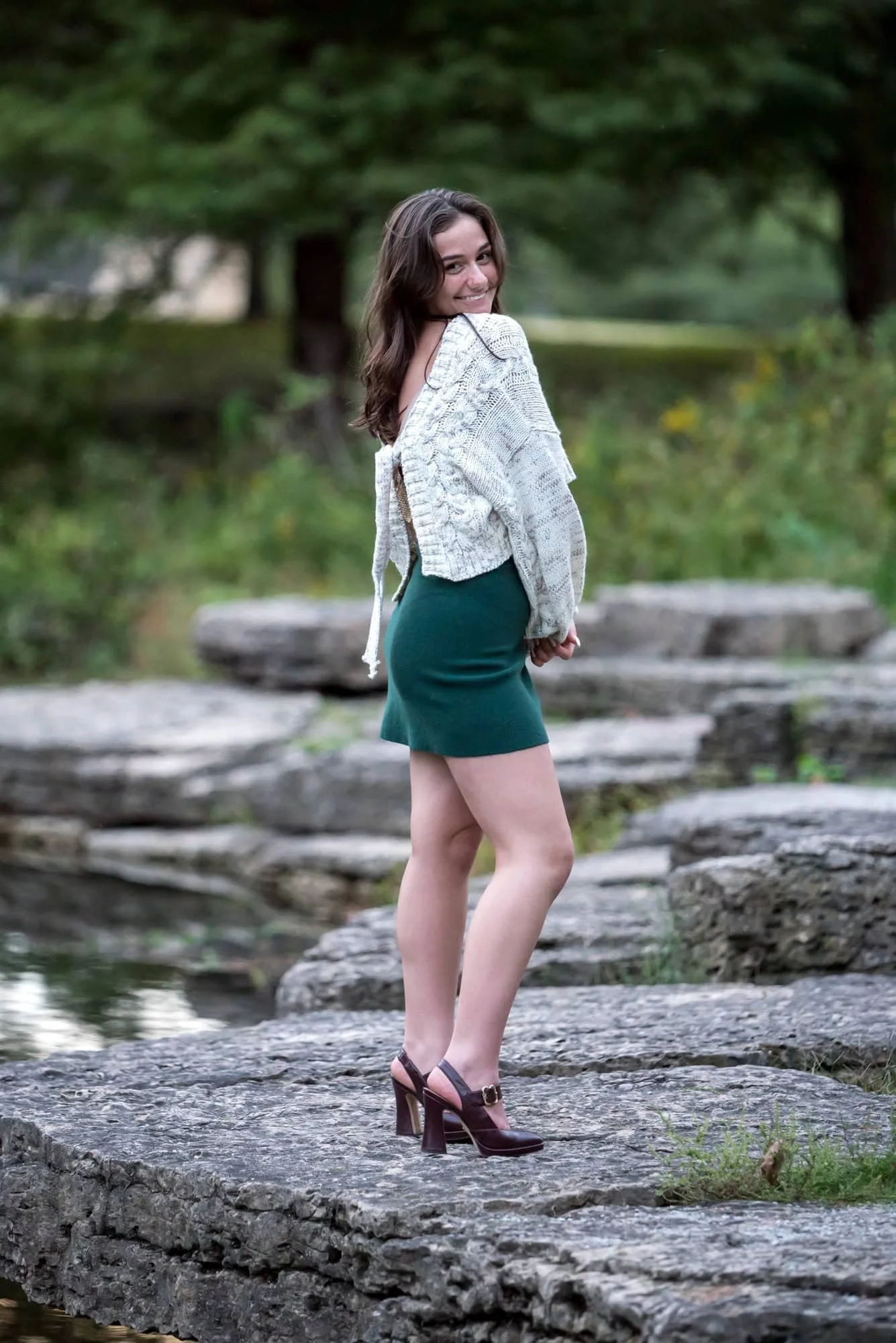 Standing on a rock near the water in St. Louis, a senior girl in a white sweater and green skirt looks back over her shoulder for a picture.