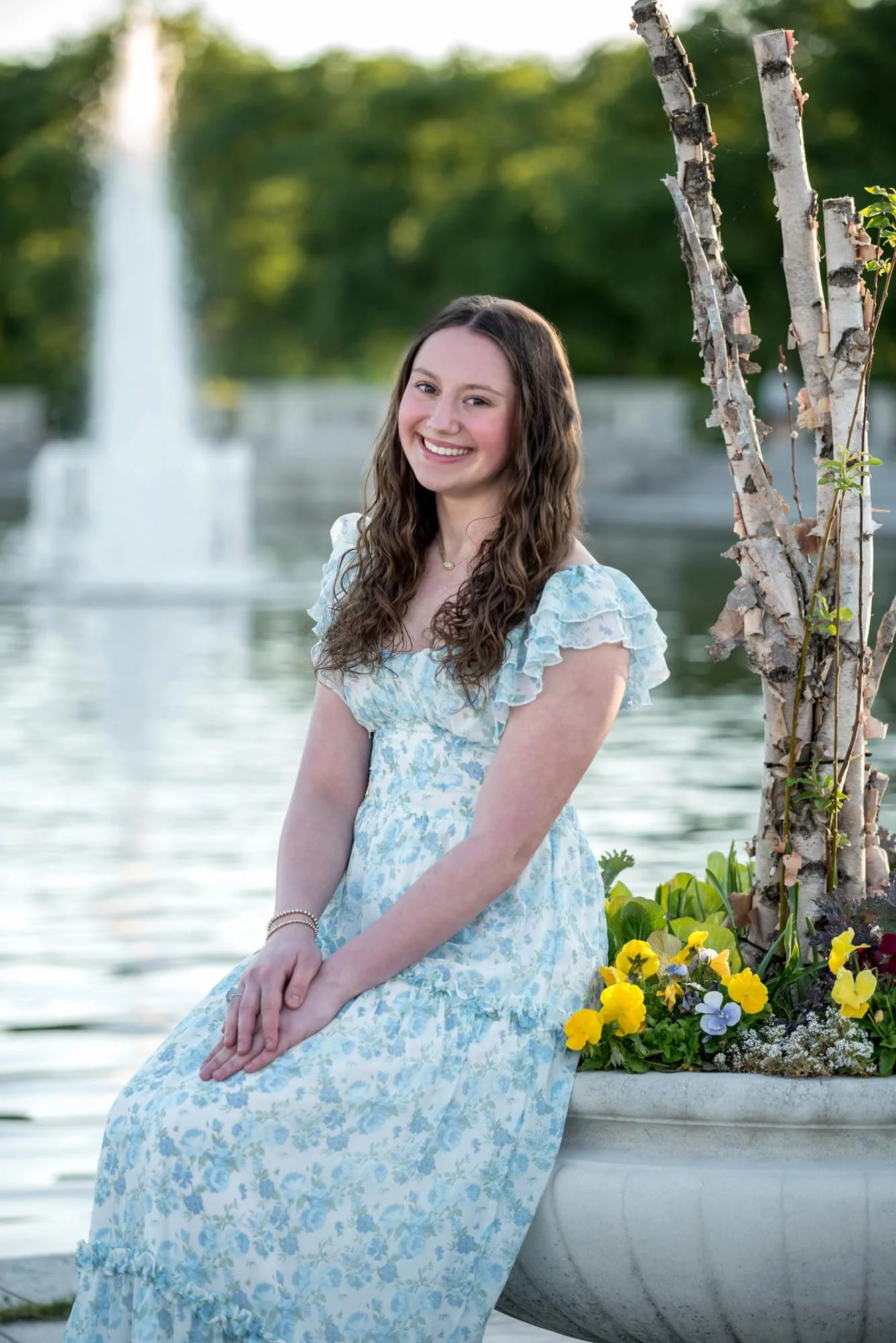 Sitting by the Grand Basin at Art Hill, a smiling high school senior girl in a light blue floral dress poses next to a flower-filled planter with a fountain blurring beautifully in the background.