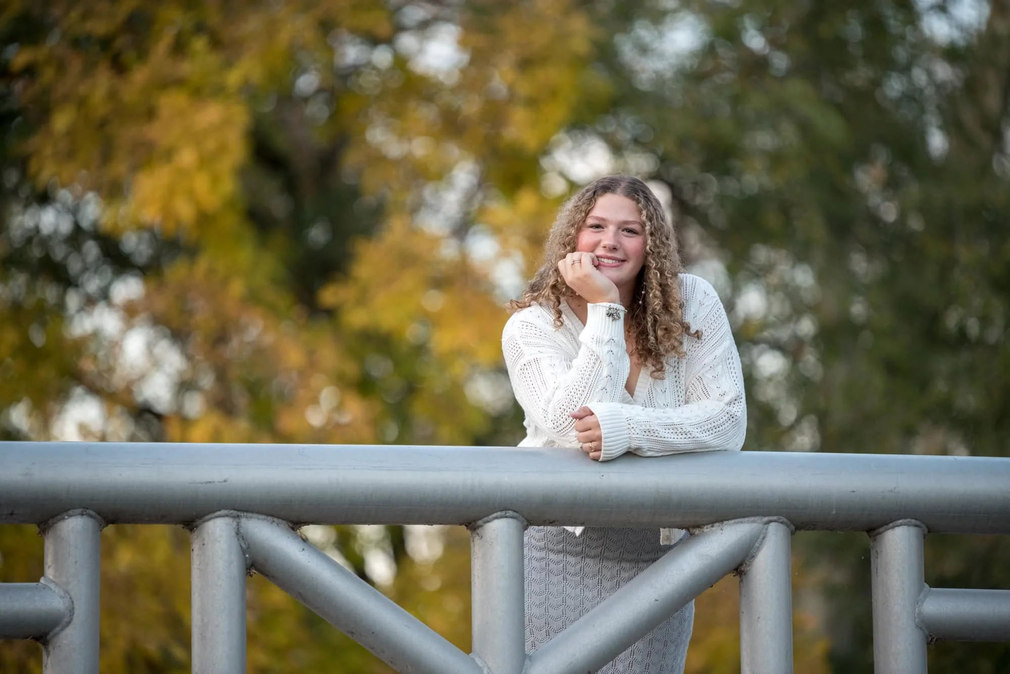 Scenic autumn senior picture capturing a girl in a white cardigan leaning on a wooden bridge railing framed by fall trees near Art Hill in Forest Park.