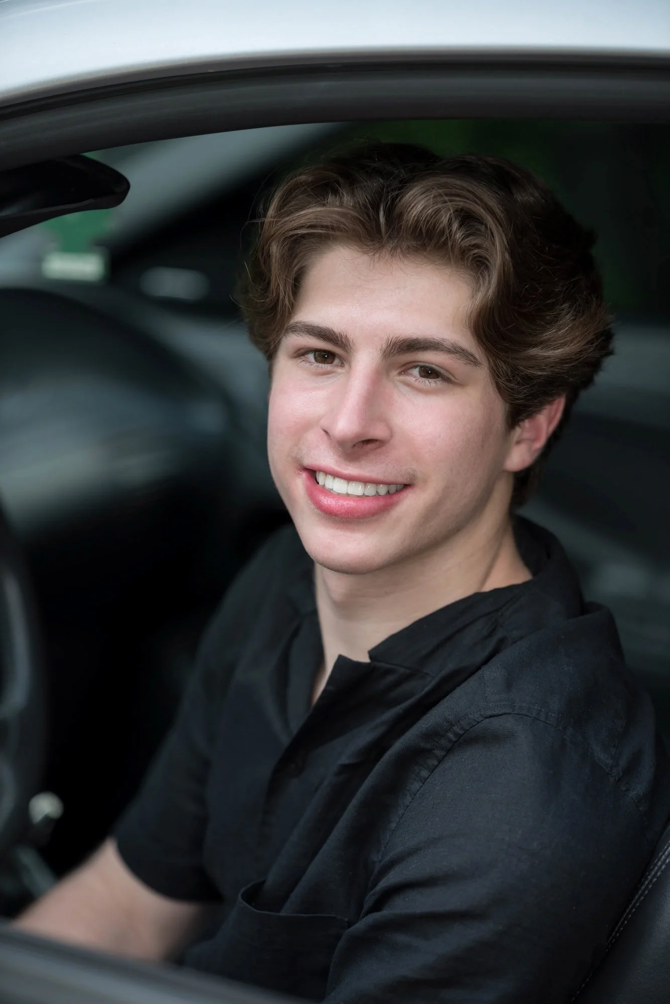 Smiling while sitting inside his car, a senior guy wearing a black shirt poses for a picture by a Columbia, IL photographer.