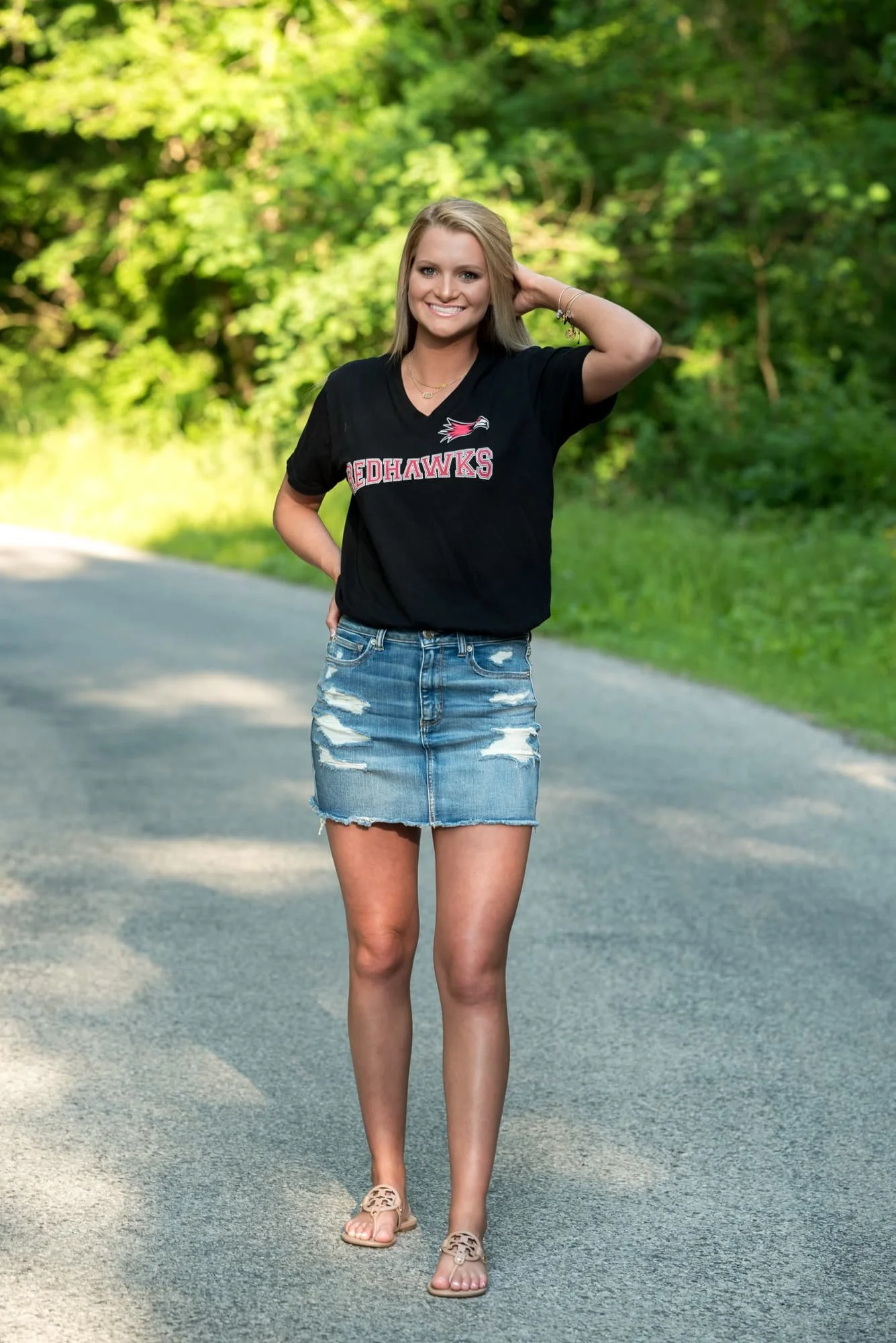 High school senior portrait by a Columbia IL senior photographer, featuring a girl standing in a sunlit roadway wearing a black Southeast Missouri State Redhawks college shirt, jean shorts, and tan flip-flops.