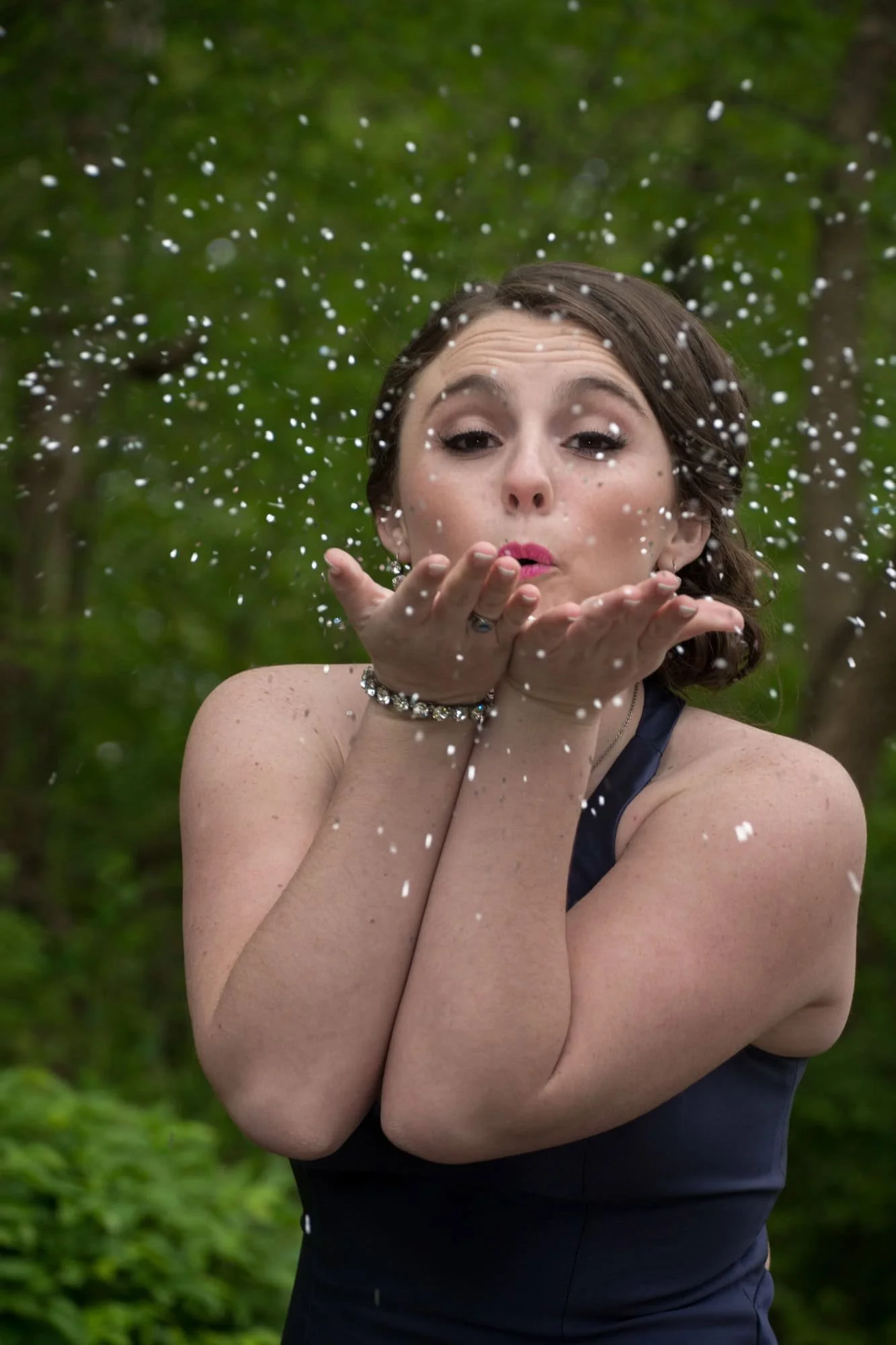 Close-up photo of a high school girl blowing silver glitter toward the camera with lush green trees blurred in the background taken by Columbia IL senior photographer.