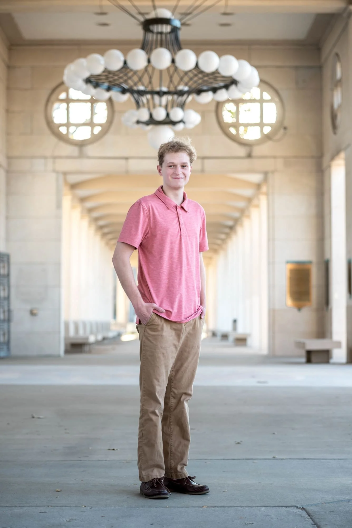 Sophisticated senior picture of a guy wearing an orange shirt, khaki pants, and brown shoes, posing with his hands in his pockets directly underneath a grand historic chandelier at The Muny in Forest Park.