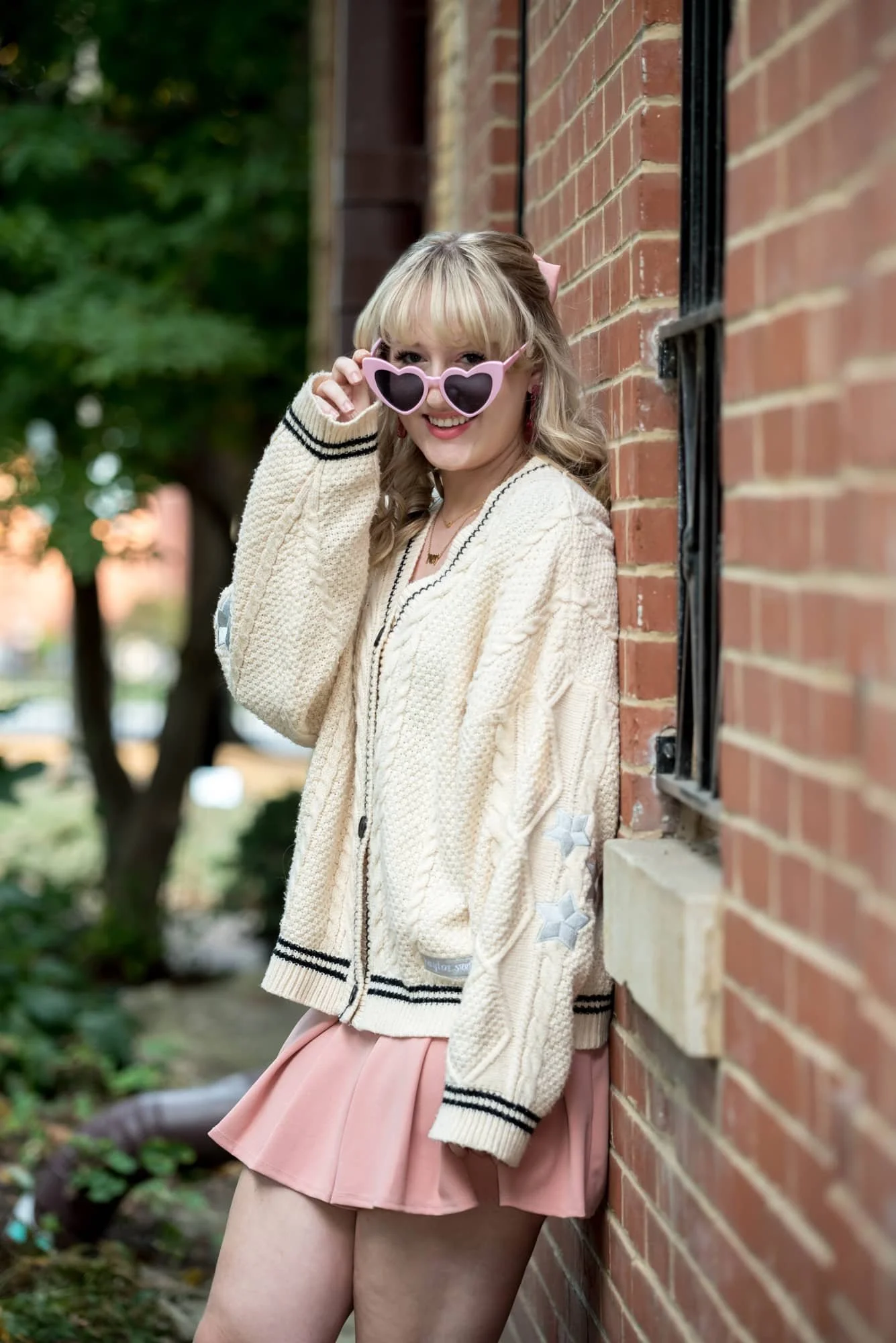 Fun and whimsical senior picture of a girl wearing pink heart sunglasses and a Taylor Swift-inspired pink skirt outfit, leaning against a historic brick building at Lafayette Park.