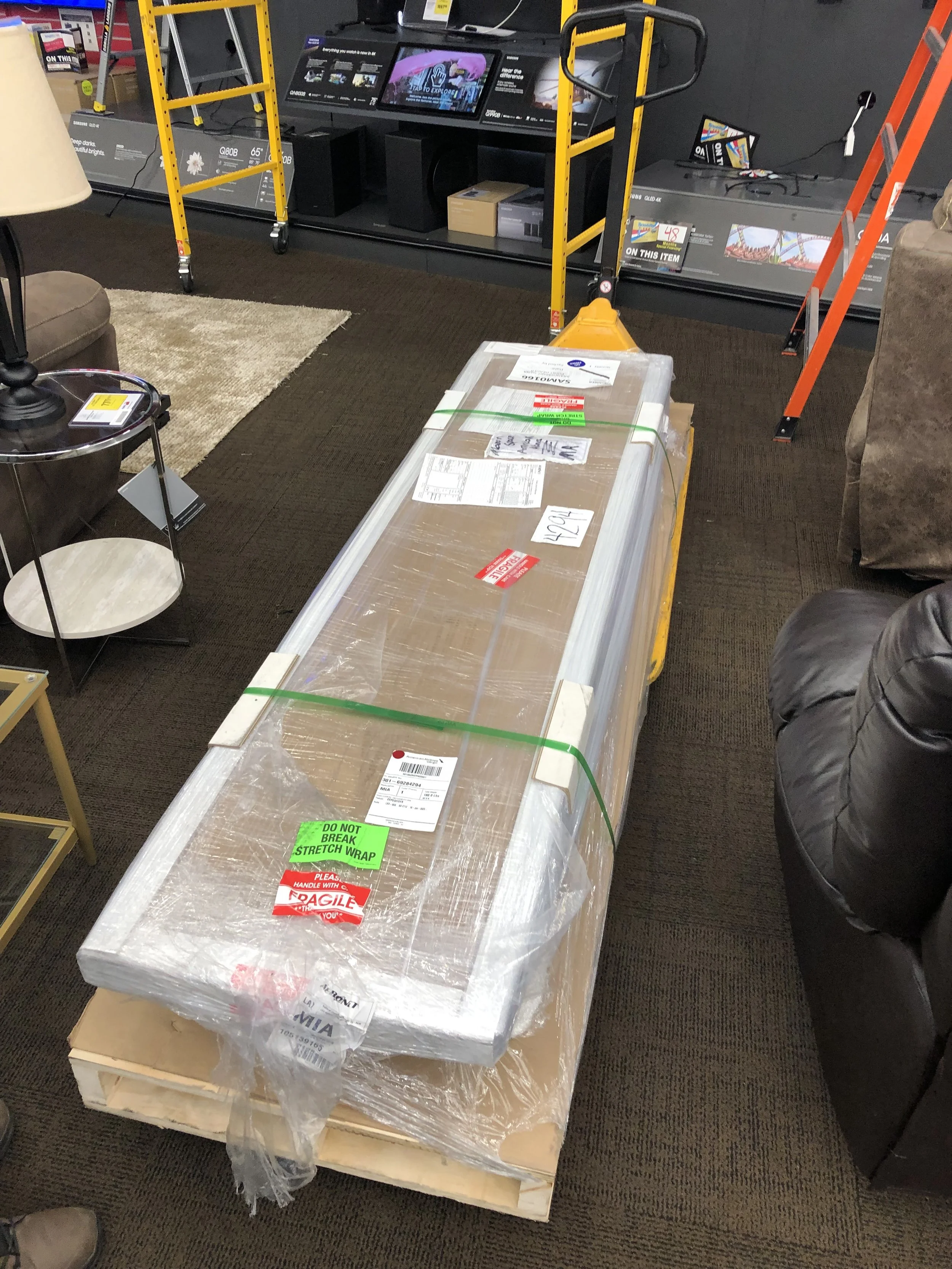 A large package wrapped in plastic sits on a wooden pallet in a store. There's a green sticker that says "Do Not Break Stretch Wrap." The package has several labels, including red "Fragile" and red "Handle with Care" stickers. To the left, there's a 