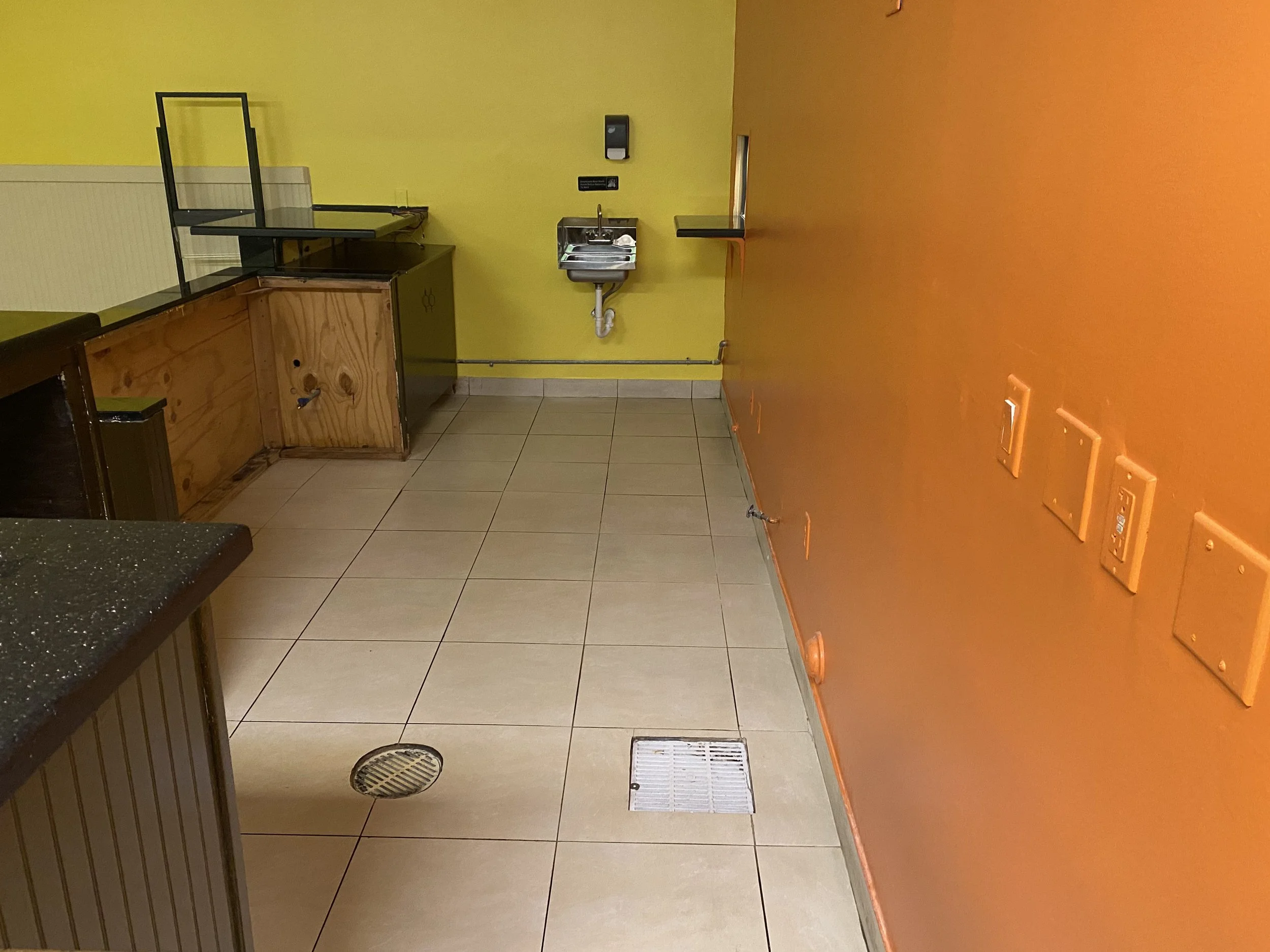 Restaurant Cleaning Service in Fort Lauderdale Kitchen or utility room with yellow and orange walls, tiled floor, stainless steel sink, wooden counter, and black metal frames, with some vent covers and a drain on the floor.