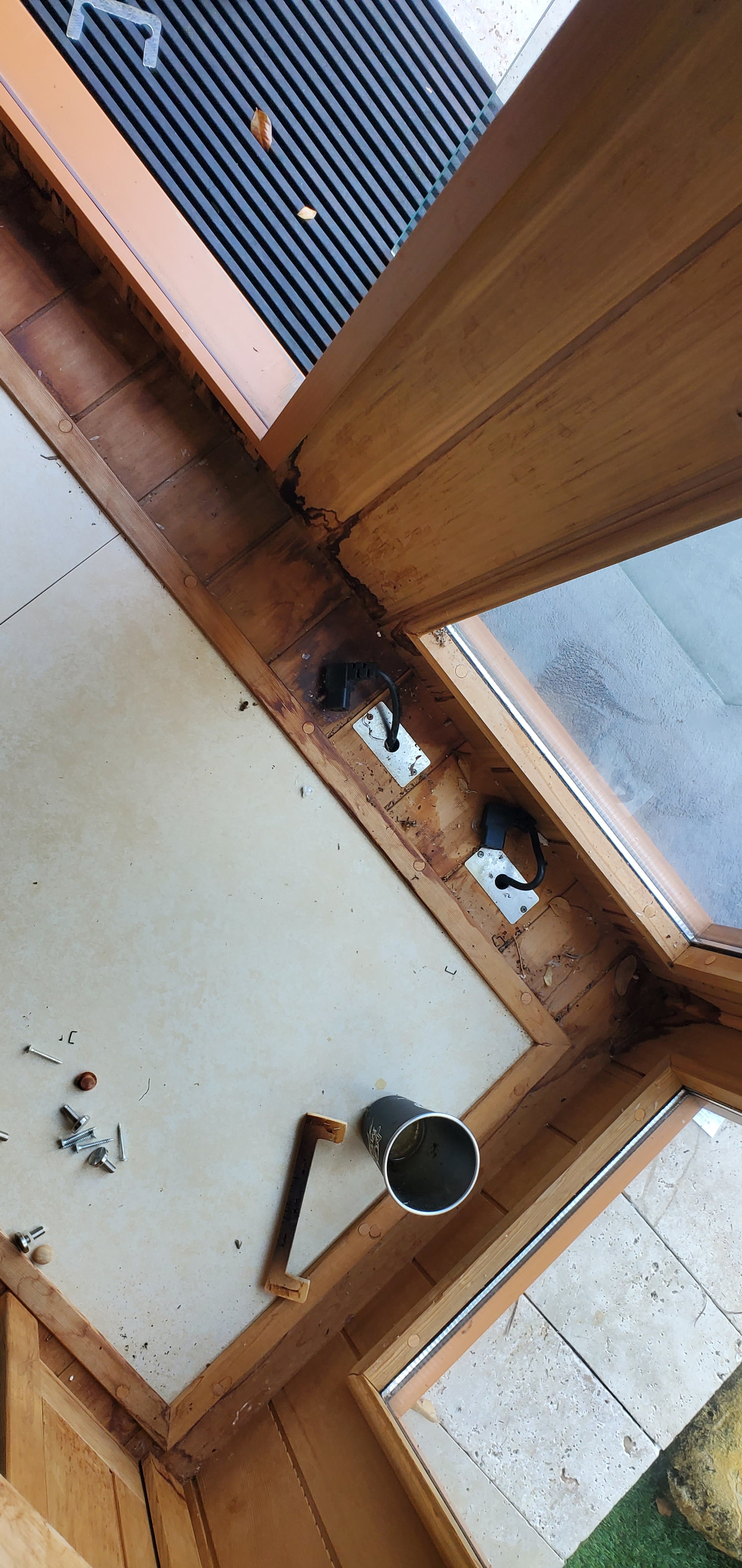Close-up of a wooden base on the floor with electrical wiring and components, including hooks and screws, near a window and stone tiles outside.