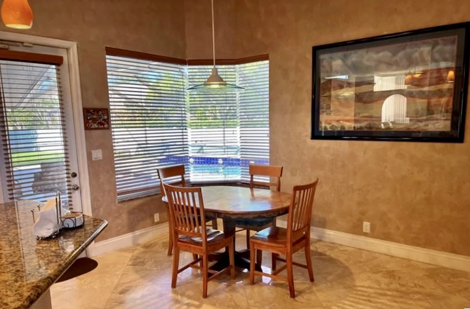 CLEANING dining area with a round wooden table and four matching chairs, large windows with blinds letting in sunlight, a hanging light fixture above, a framed picture on the beige wall, a door with blinds, and a granite countertop with a tissue box.