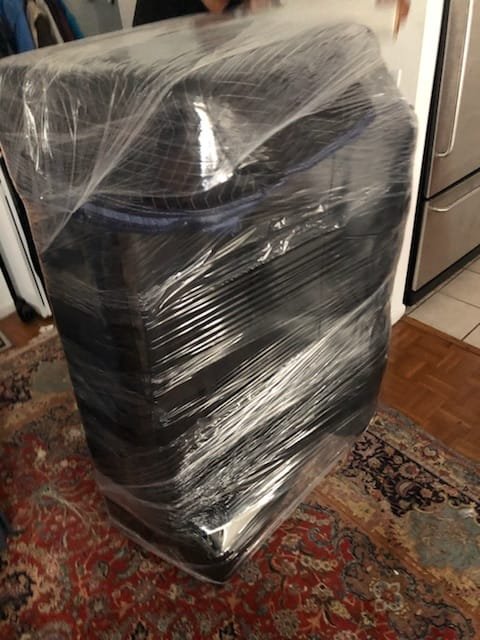 Moving Services A washing machine wrapped in clear plastic wrap, placed on a patterned rug in a kitchen.