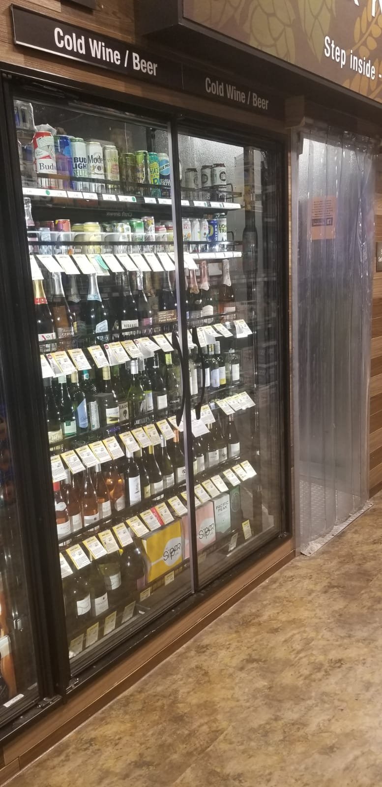 Glass-door beverage cooler filled with assorted bottles of wine and canned drinks, with signs indicating it contains cold wine or beer.