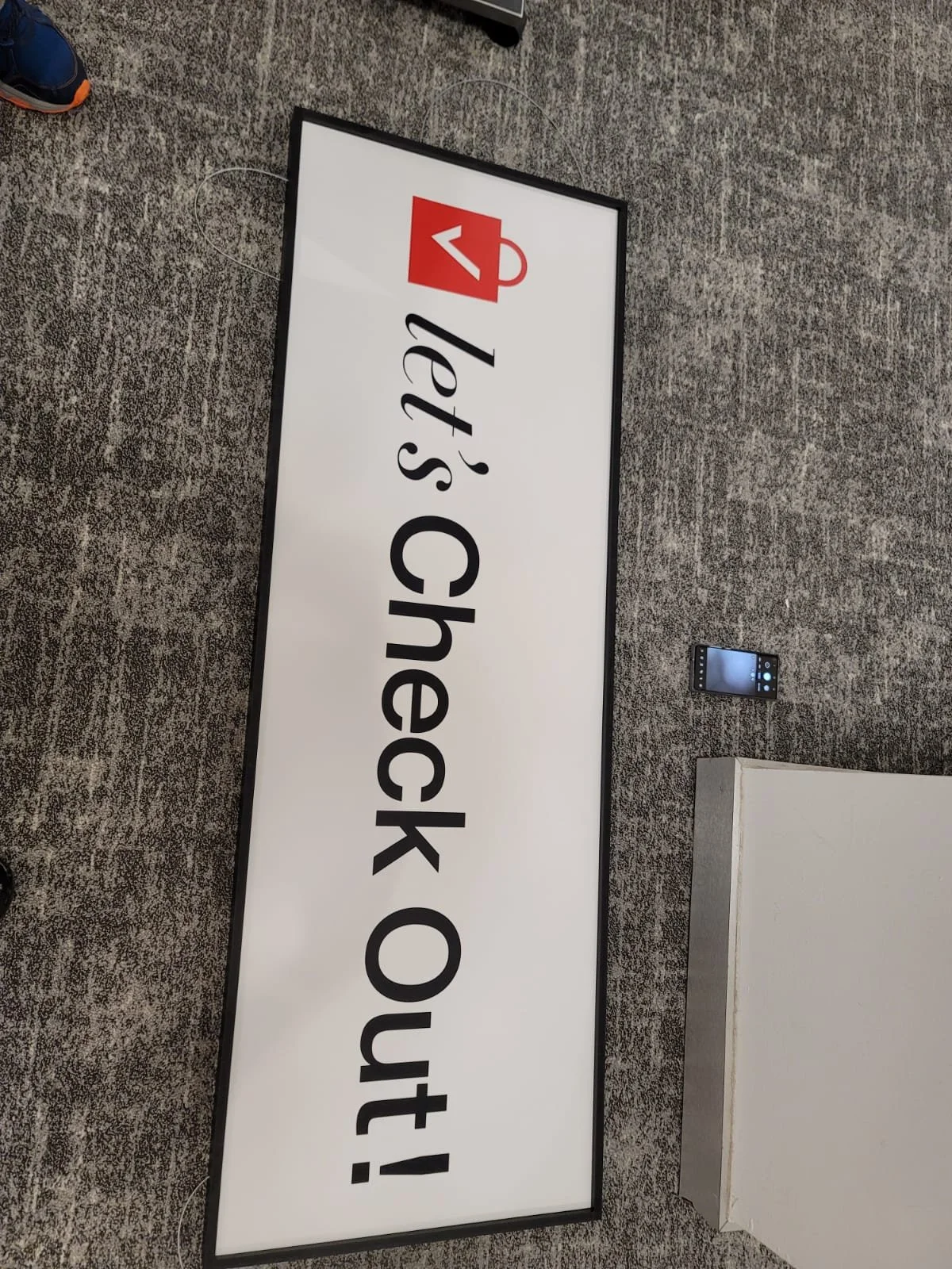 A digital sign with the text 'Let's Chat about!' and an icon of a red coffee mug with a heart design on it. A smartphone is placed on the carpet next to the sign, and the background features a textured gray carpet.