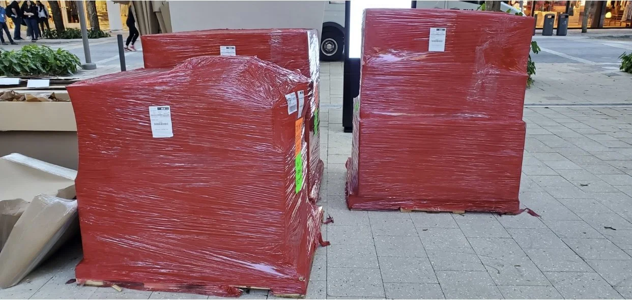 Pick Up and DeliveryLarge red pallets wrapped in plastic film placed on a sidewalk at a retail store.
