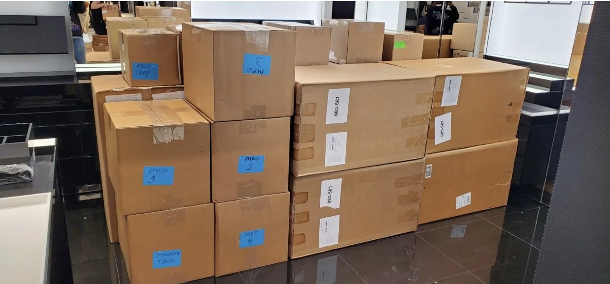 MOVING Service,  A stack of cardboard boxes labeled with blue and white stickers, located in a retail or storage area with a black tiled floor and reflective surfaces.