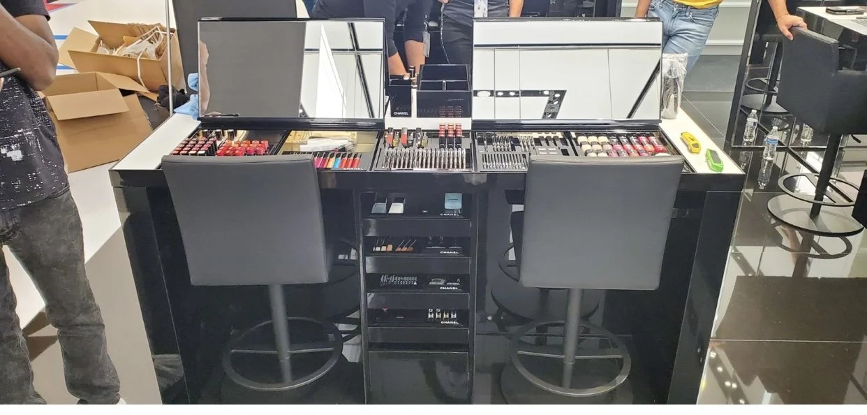 cleaning service, A display table with makeup products, including lipsticks and brushes, in a retail store. Two black chairs are in front of the table, and several people are visible in the background.