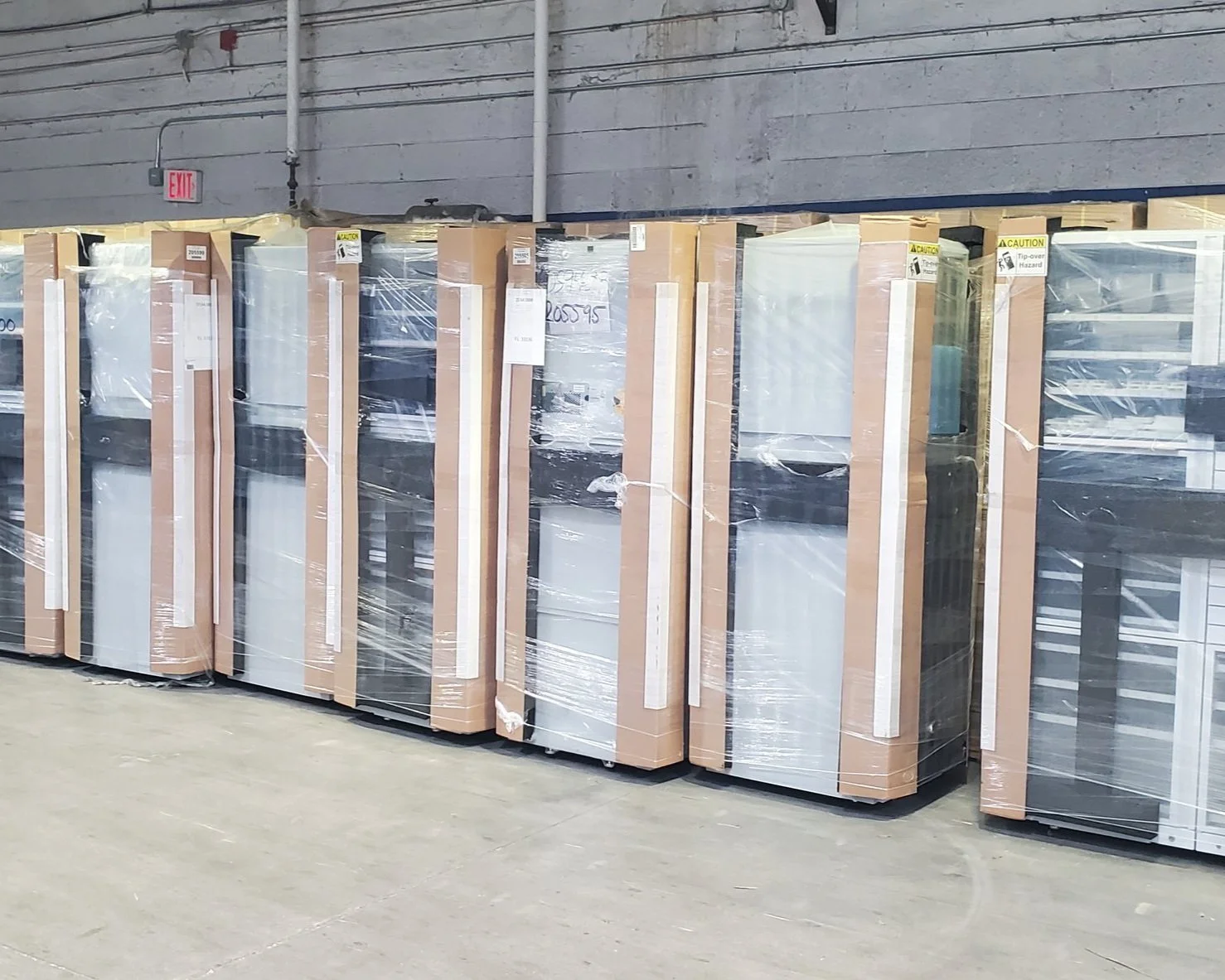 unloading load service, A row of refrigerators wrapped in protective plastic and cardboard in a warehouse or store, with a gray wall and exit sign in the background.