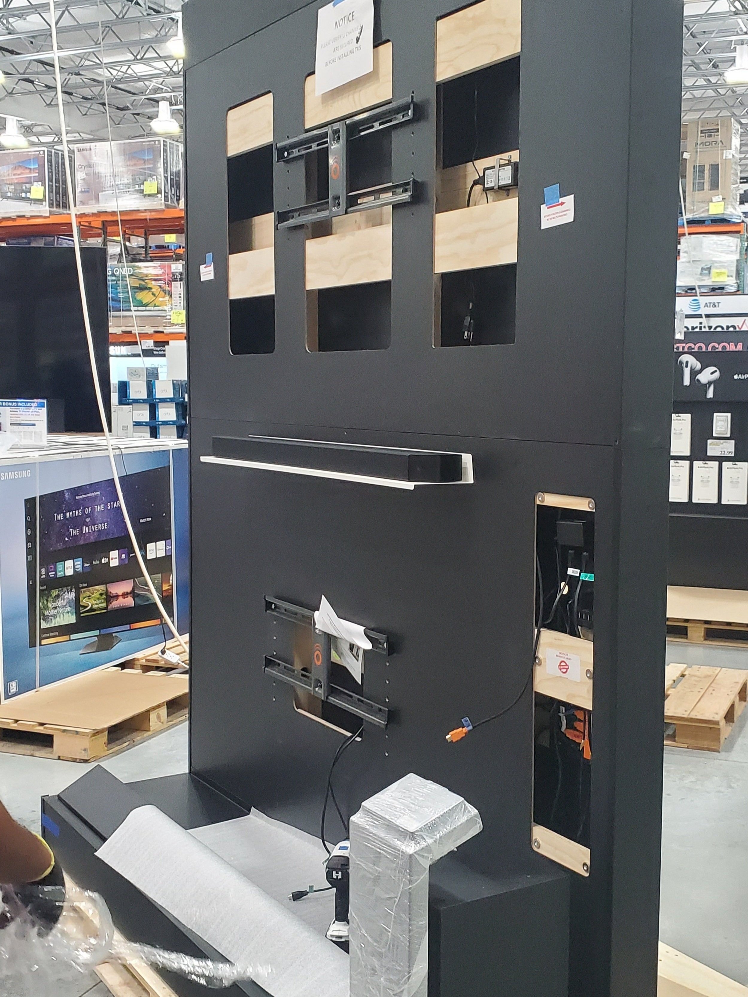 Best Installation Service in  West Palm Beach commercial office Black display unit with wooden panels and metal brackets, showing an empty slot for a television or monitor, located in a store aisle with shelves and boxes in the background.