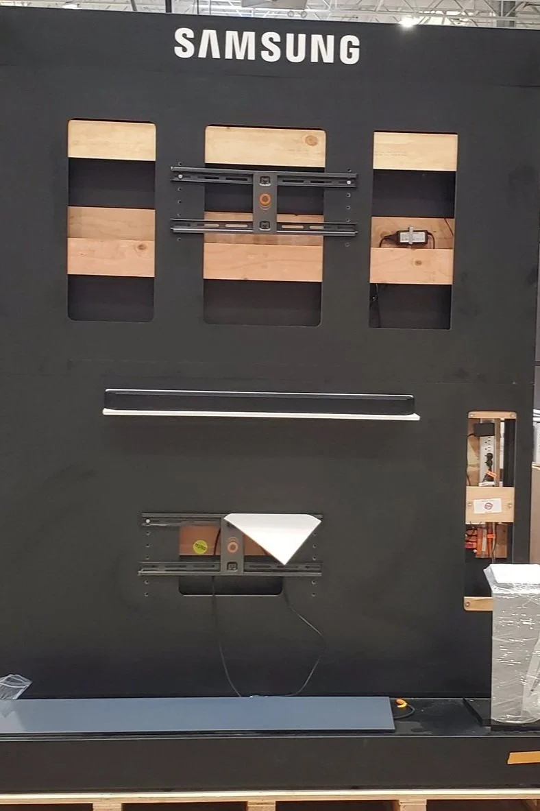 Unassembled Samsung commercial display or monitor frame in a store, with mounting hardware and brackets attached, and a wooden back panel.