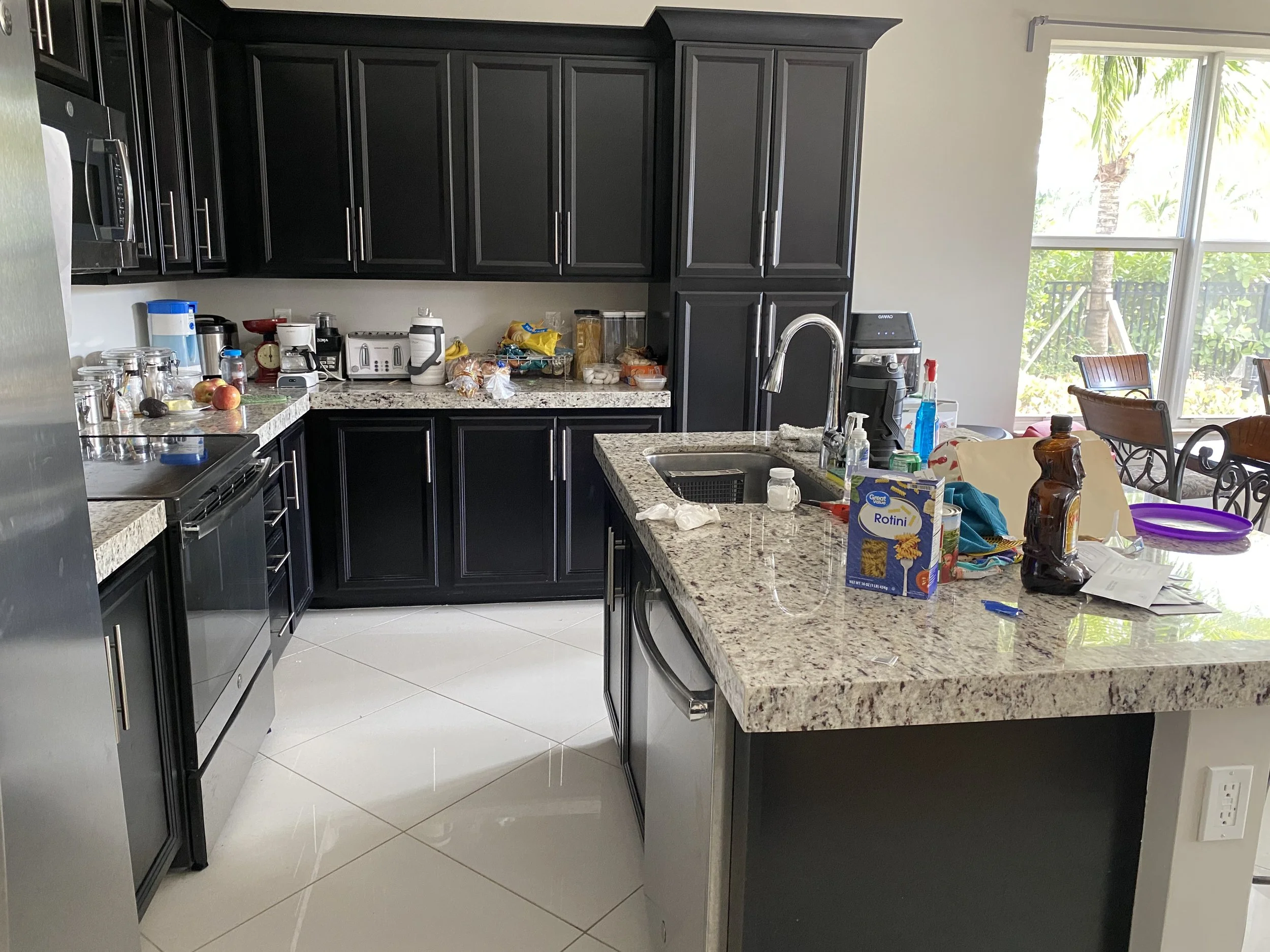 A kitchen with black cabinets, granite countertops, and a large window overlooking trees. The countertop has various items, including apples, a scale, a toaster, and snacks. The kitchen island has a sink and a bottle of cleaning spray.