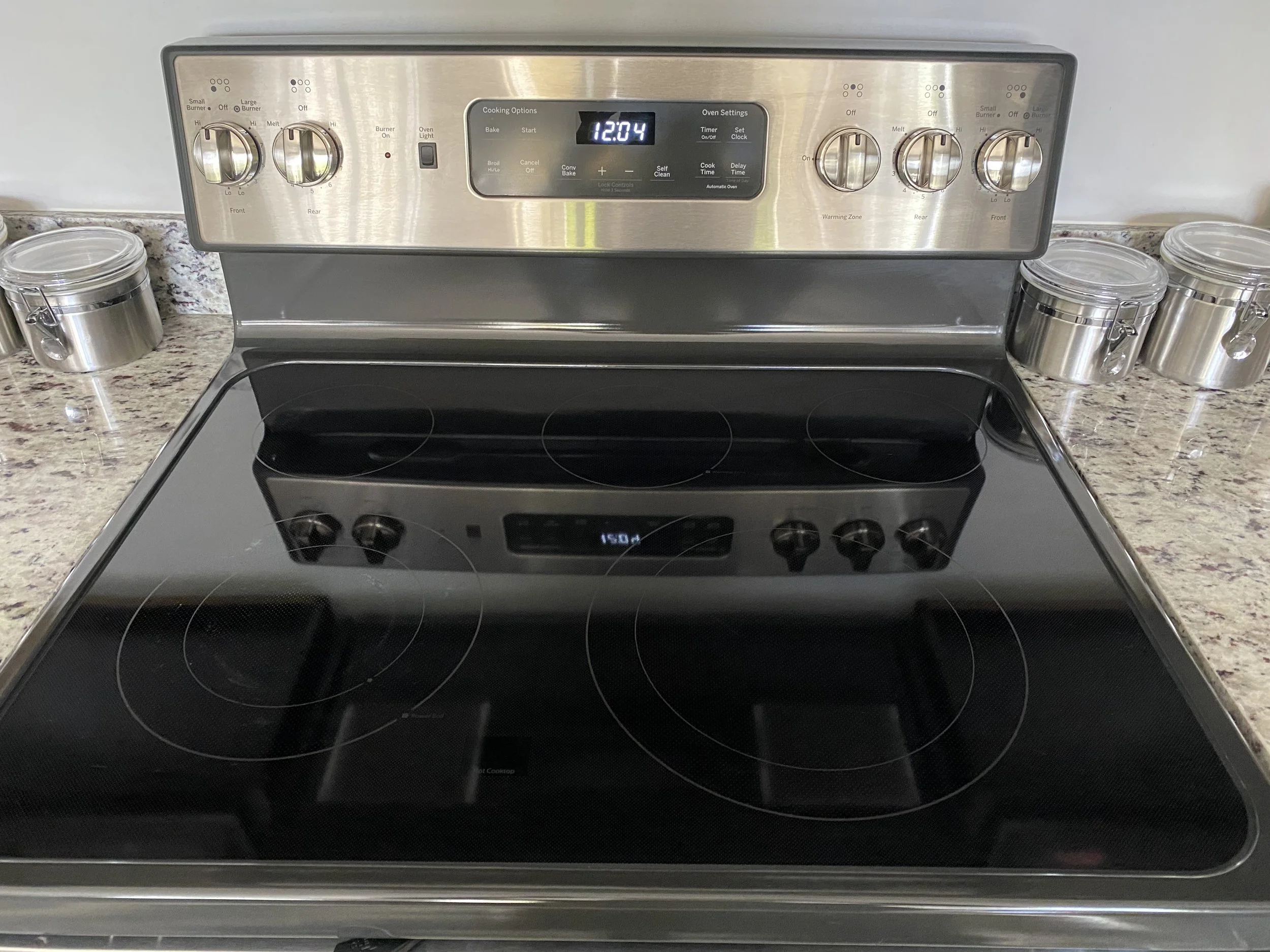 A modern stainless steel electric stove with a black glass cooktop and a digital clock showing 12:04. The stove is situated on a speckled granite countertop with three small clear storage jars with metal clips on the left and right sides.