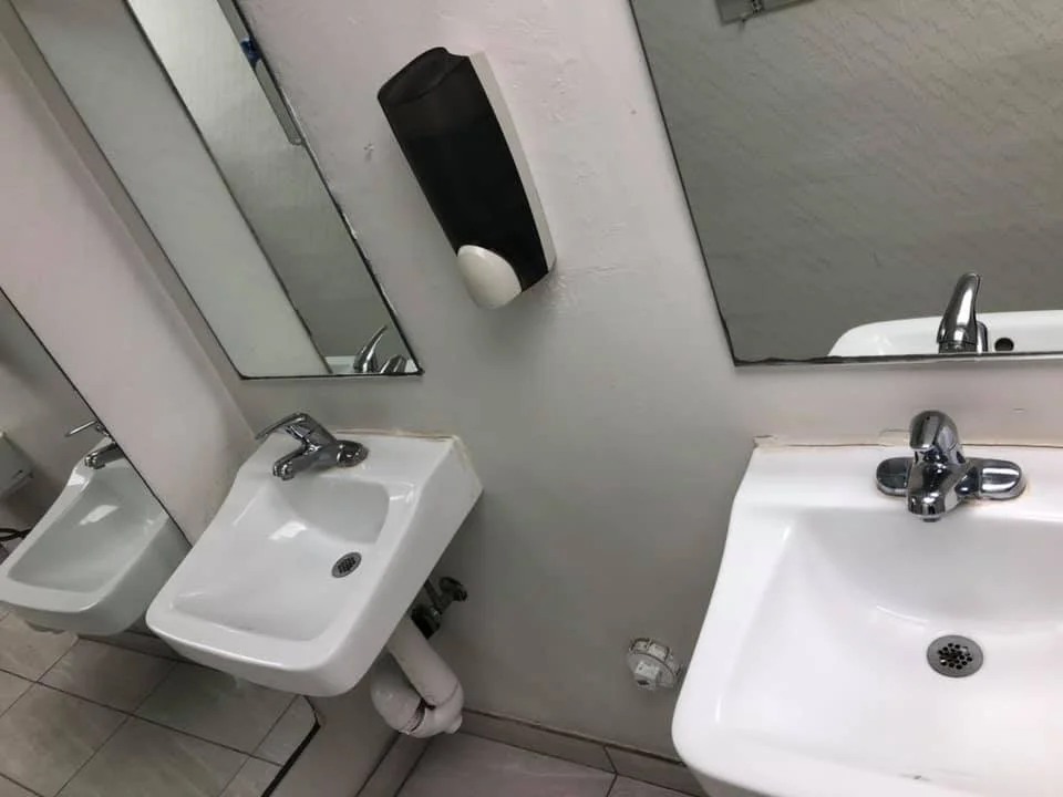 Two white sinks with silver faucets, a mirror, and a black soap dispenser on a white wall in a public restroom.