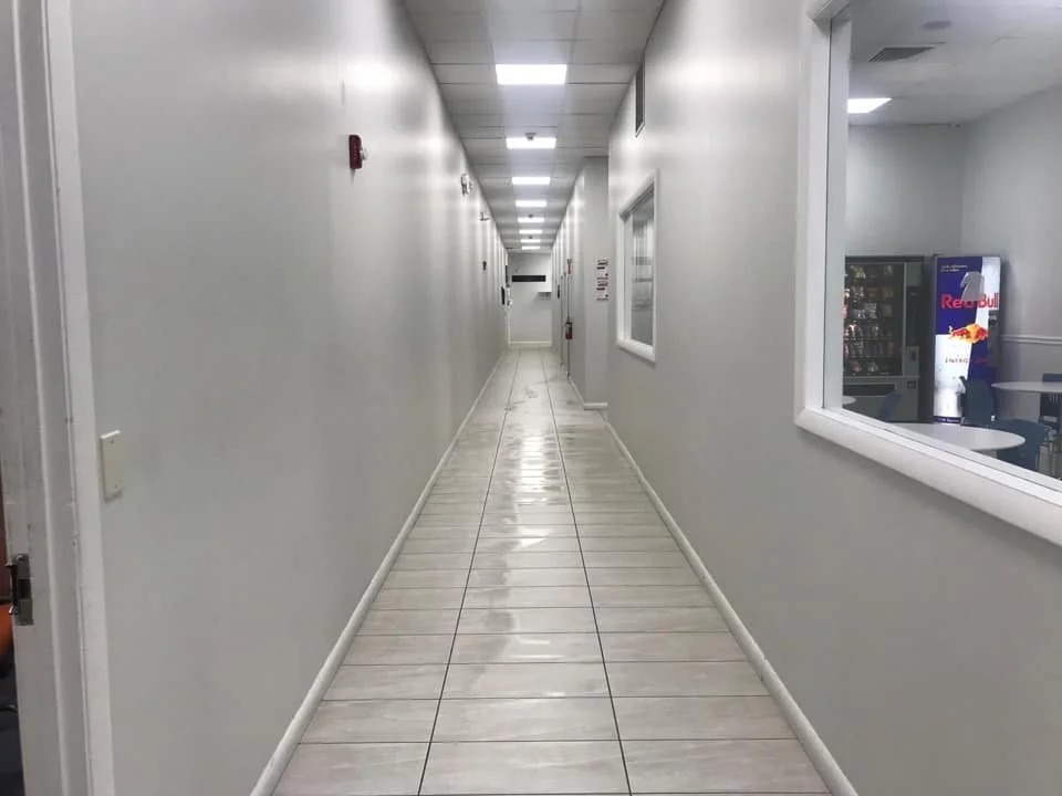 commercial cleaning in Fort Lauderdale, Empty hallway with white walls, tiled floor, and ceiling lights, and a window showing a vending machine and Red Bull fridge.