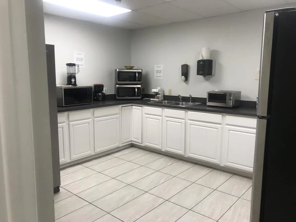 office cleaning service, Empty kitchen with white cabinets, black countertops, and beige tiled floor, featuring appliances like a refrigerator, microwave ovens, coffee maker, blender, toaster oven, and a double sink.