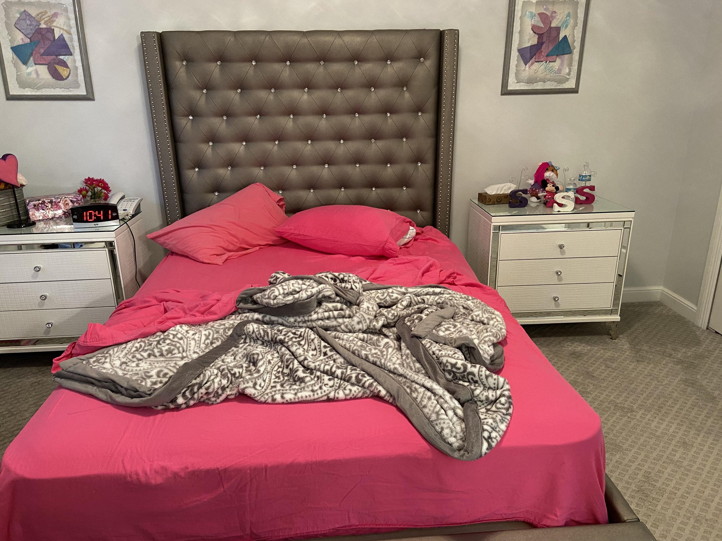 Unmade bed with pink sheets and a gray blanket on top, in a bedroom with white nightstands on each side. The left nightstand has a small lamp, flowers, a phone, and a digital clock displaying 10:41. The right nightstand has plush toys, decorative let