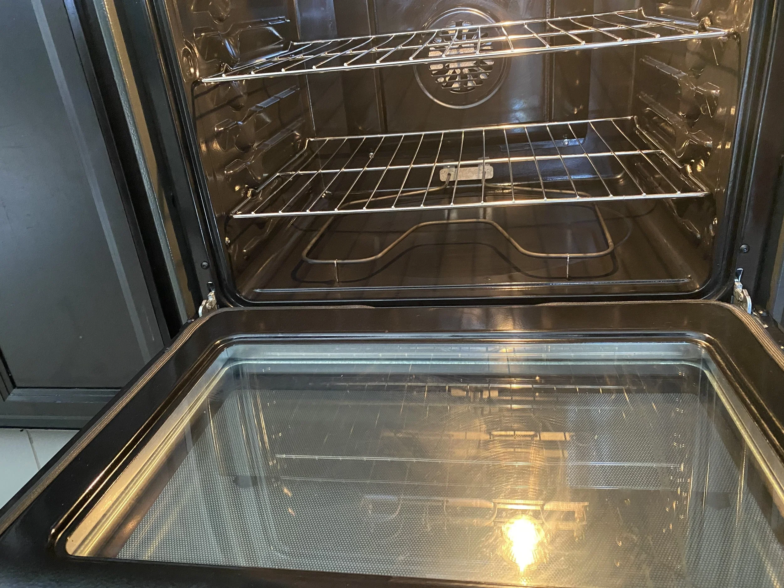Empty stainless steel oven with two oven racks, near open oven door, reflecting light.
