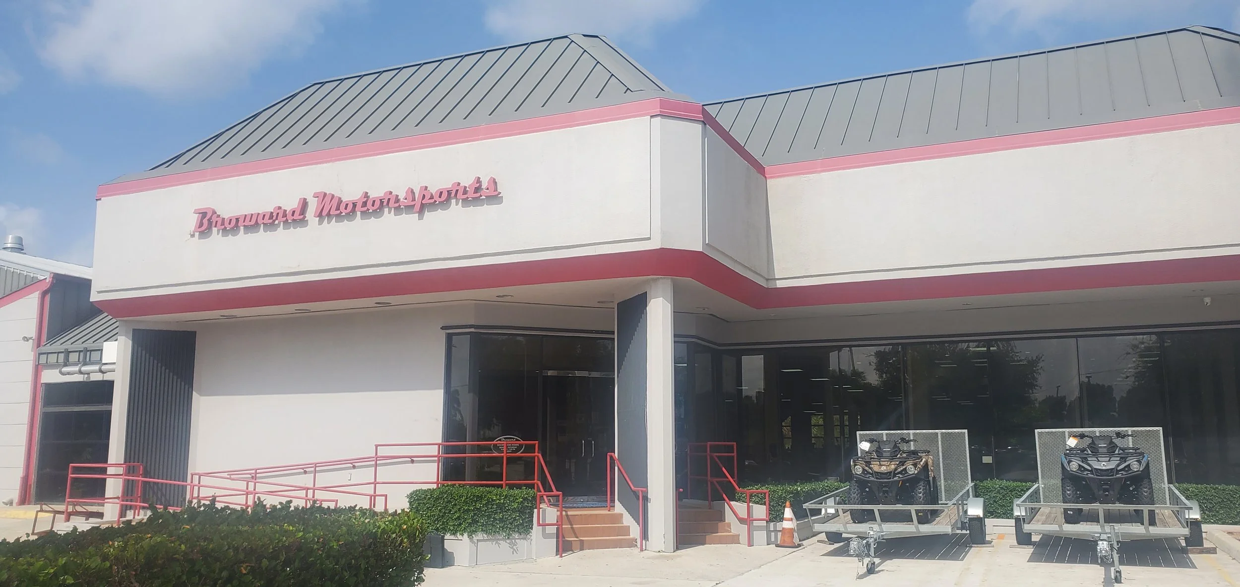 Exterior view of Broward Motor Sports building with a white facade, pink trim, and a gray metal roof. There are motorcycles parked in front and red railings leading to the entrance.