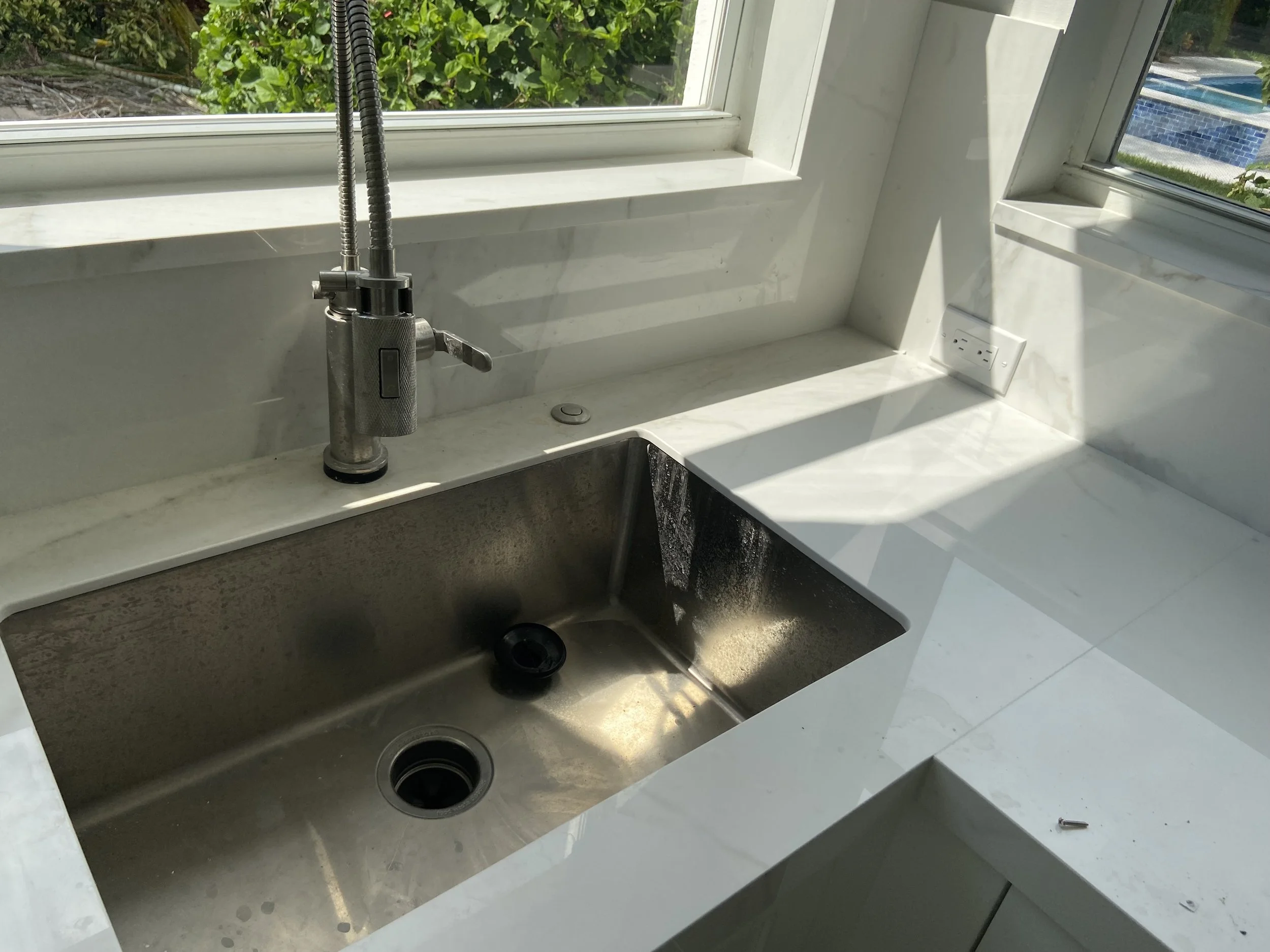 Modern kitchen with a white marble countertop, stainless steel sink, tall faucet, and an electrical outlet on the wall near the window. Outside, green trees, a backyard, and a swimming pool are visible through the window.