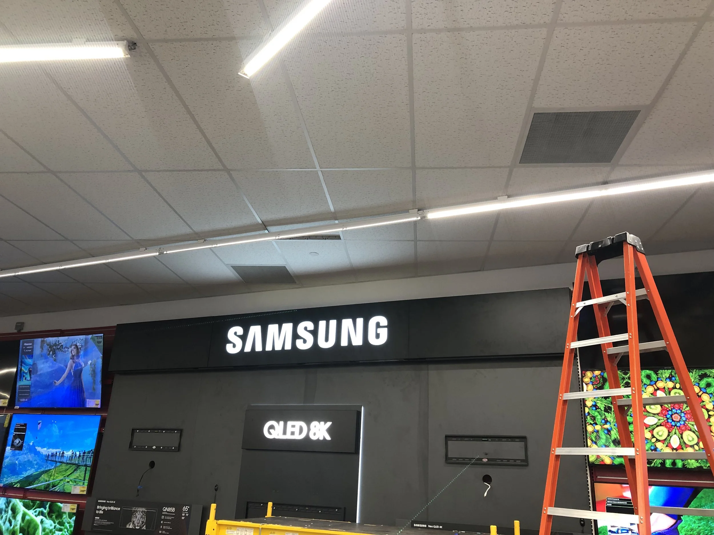 Looking up at the ceiling of an electronics store, with a bright light fixture, a vent, and part of a ladder visible, along with a store sign reading "SAMSUNG."