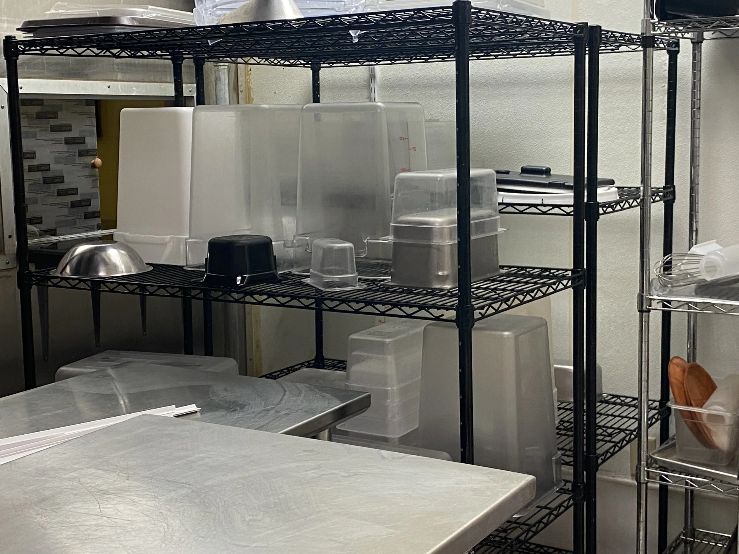 best restaurant cleaning service, A kitchen or storage room with black metal shelving units holding various plastic containers and kitchen supplies, with a stainless steel countertop in the foreground.