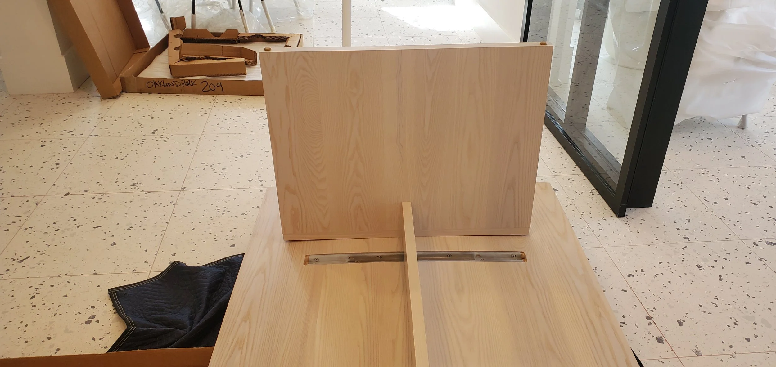 Wooden furniture pieces, including a large panel and a smaller slab, are in a room with a tiled floor. A black quilted item is on the floor, and another piece of wood with a metal rail is visible. There is a glass door and cardboard boxes in the back