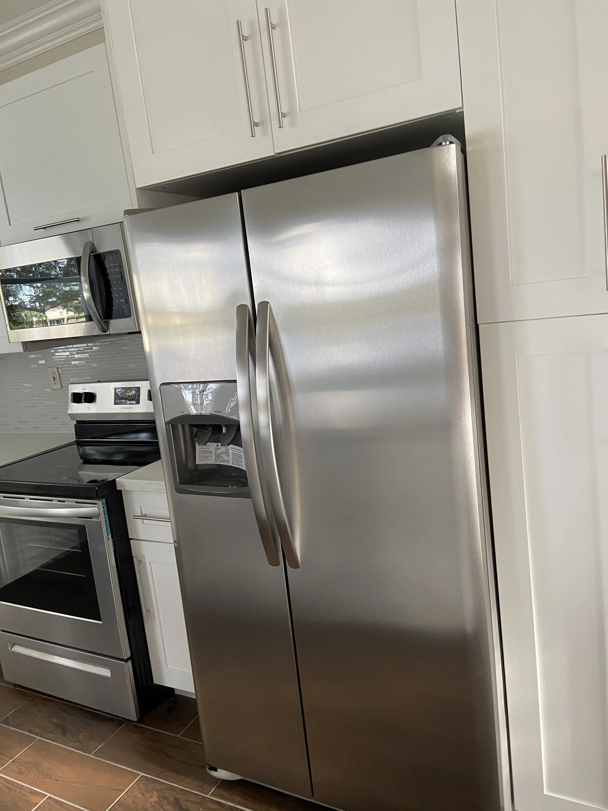 Stainless steel side-by-side refrigerator in a modern kitchen with white cabinets and brown tiled floor.