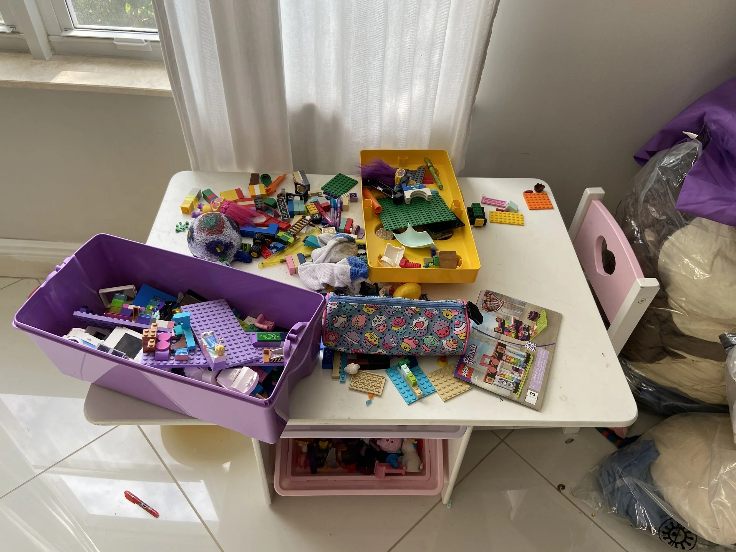 Residential house cleaning in Fort Lauderdale A white table filled with scattered LEGO pieces, containers, and toys, with a window and curtains in the background.