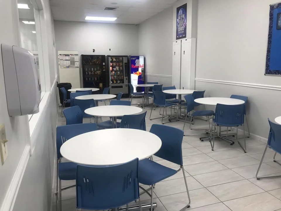 Clean break room with white walls, white tile floor, several white round tables, and blue chairs. Vending machines and a bulletin board are in the background.