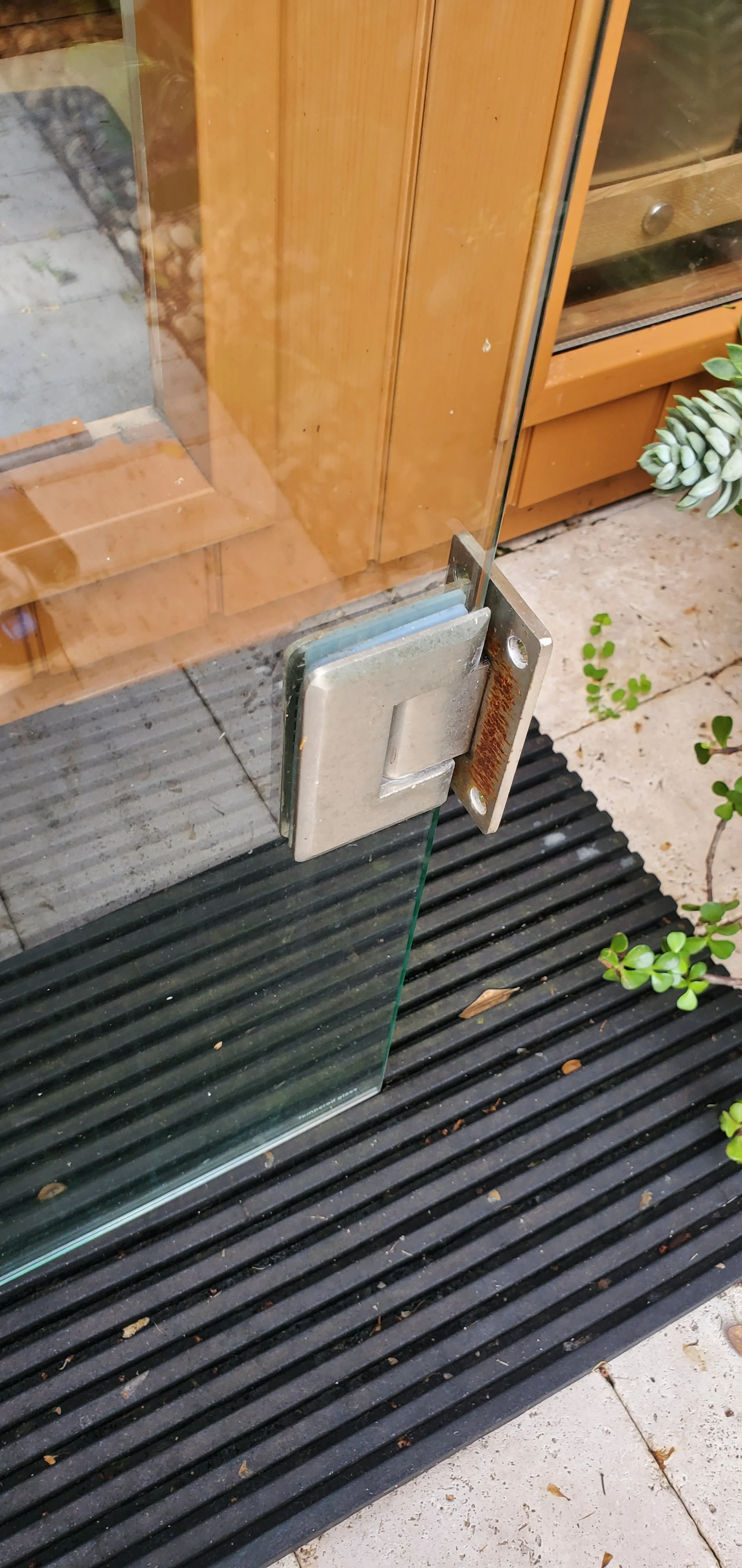 Installation Service in Fort Lauderdale Close-up of a glass door with a metal latch, showing rust on the latch and a black rubber mat outside with small plants nearby.