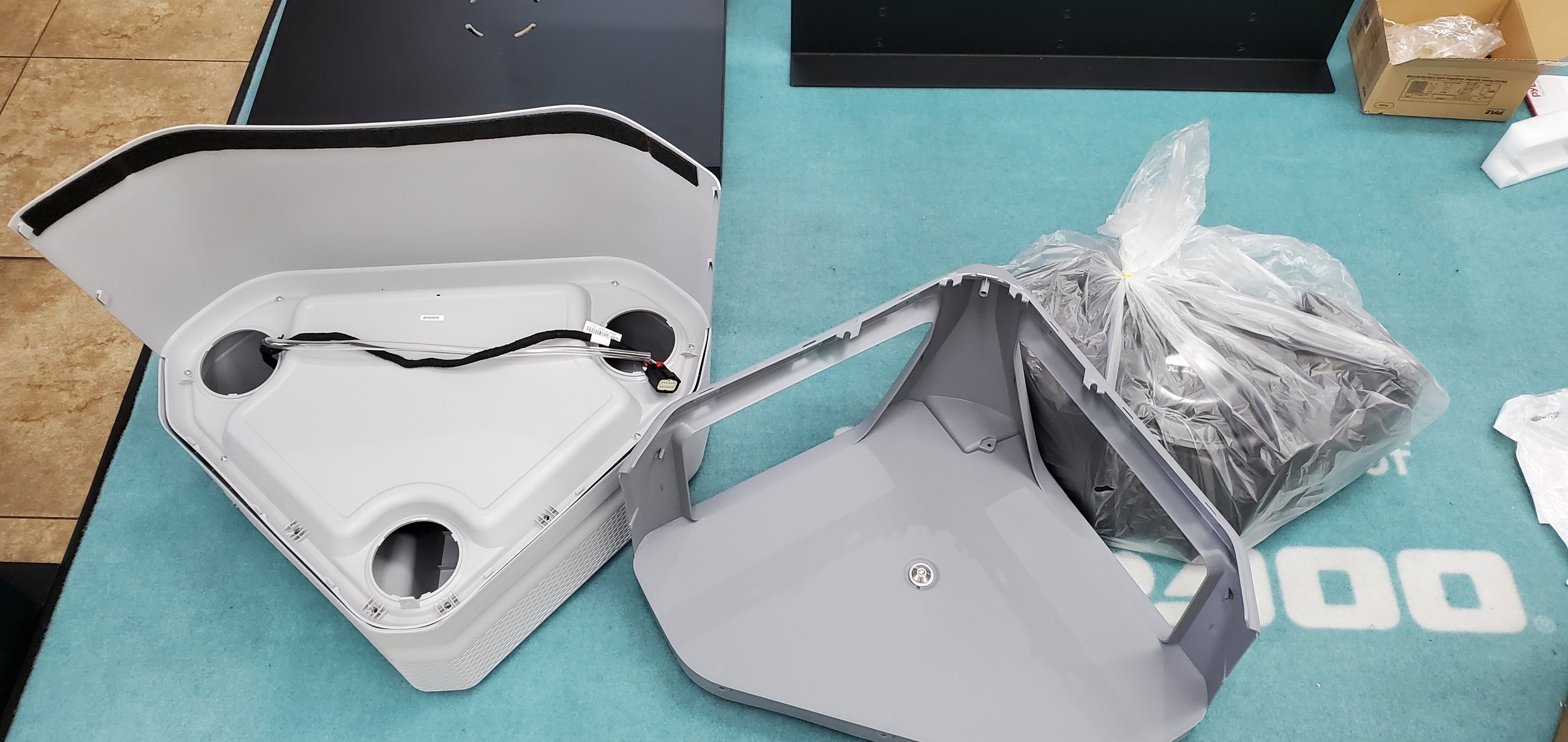 Disassembled electronic device parts on a turquoise work surface, including a white plastic shell, cables, and a wrapped component in plastic.